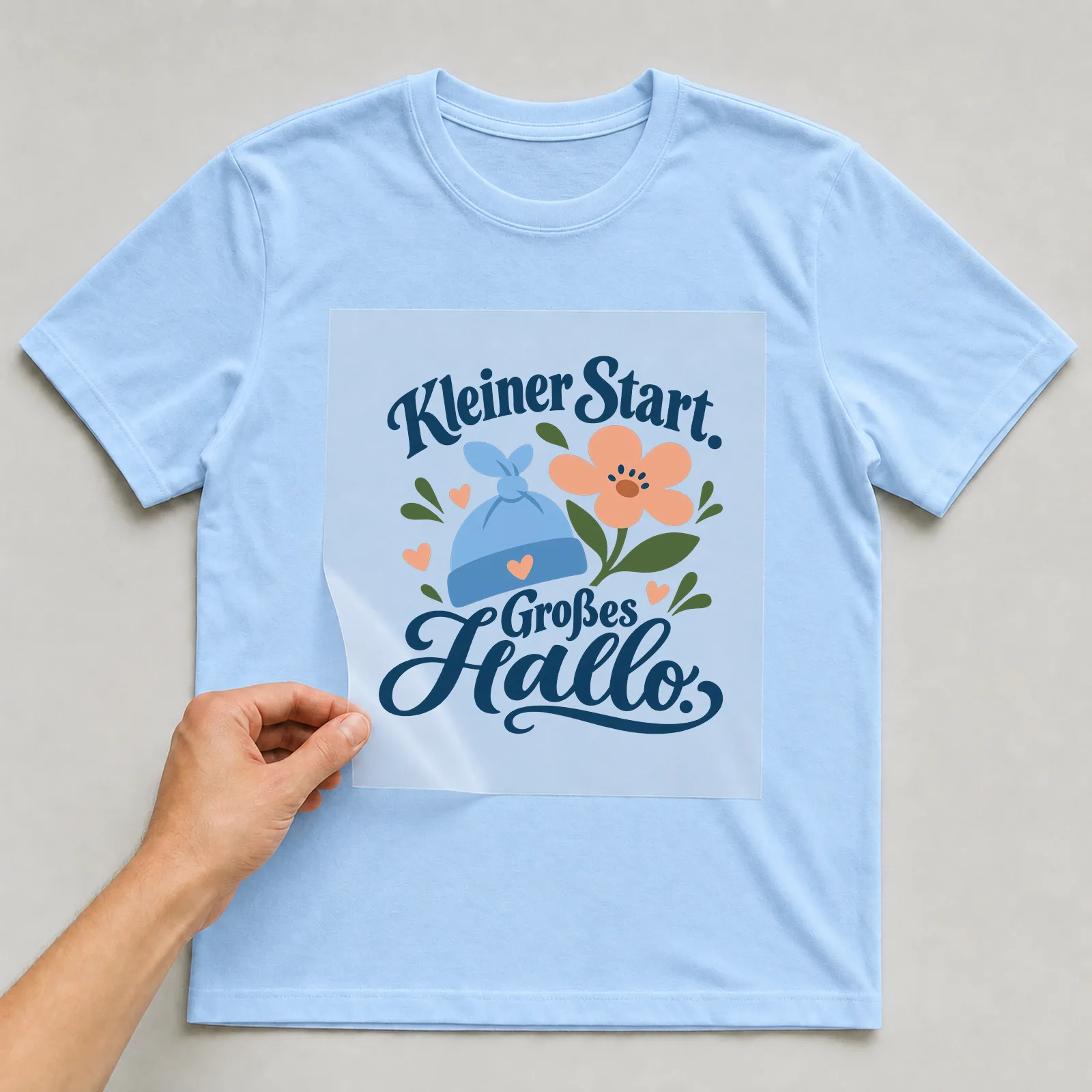 Bügelbild „Kleiner Start. Großes Hallo.“ auf hellblauem T-Shirt, Folie wird aufgelegt