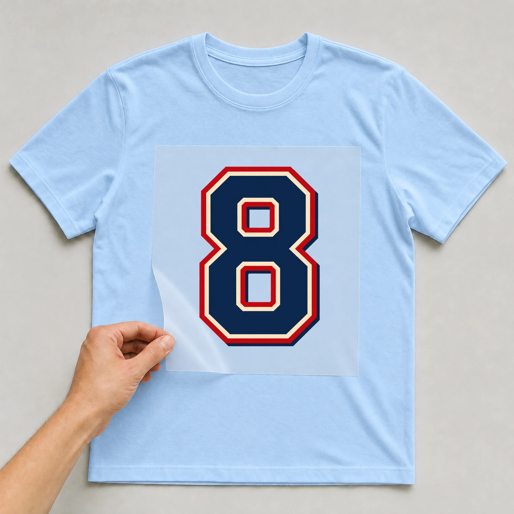 Bügelbild „Zahl 8 im Retro-College-Stil“ auf babyblauem T-Shirt, Folie wird aufgelegt