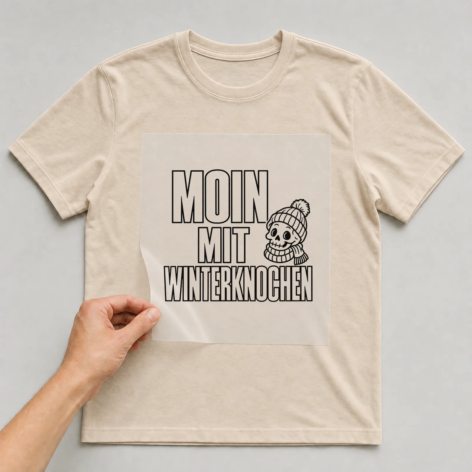 Bügelbild „Moin mit Winterknochen“ auf beigem T-Shirt, Folie wird aufgelegt