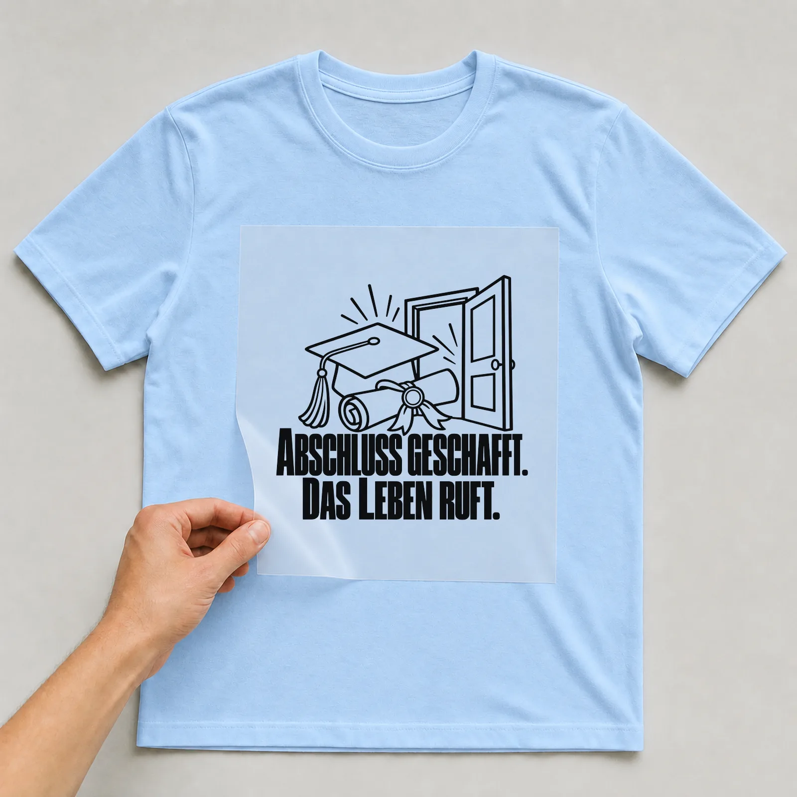 Bügelbild „Abschluss geschafft. Das Leben ruft.“ auf hellblauem T-Shirt, Folie wird aufgelegt