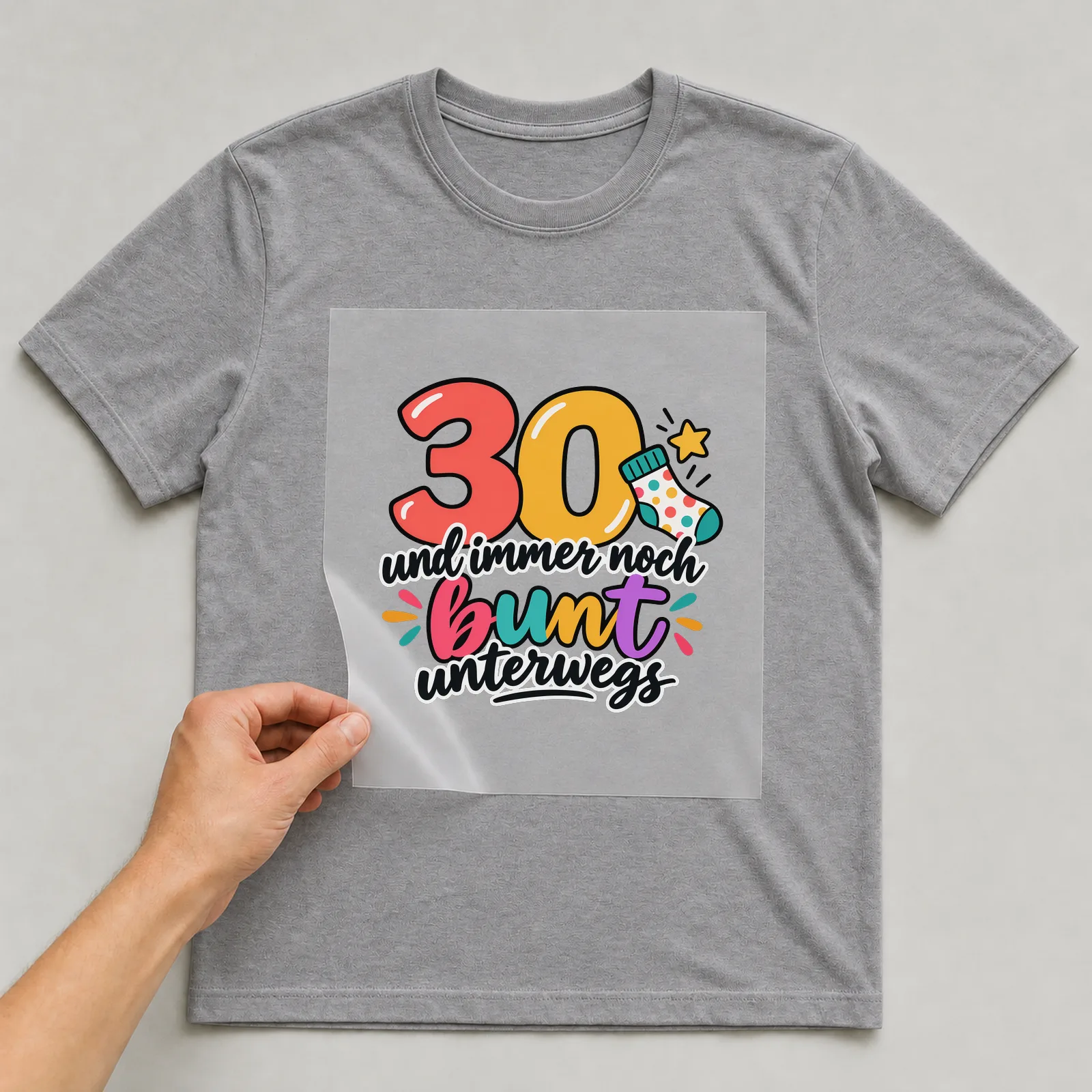 Bügelbild „30 und immer noch bunt unterwegs“ auf grauem T-Shirt, Folie wird aufgelegt