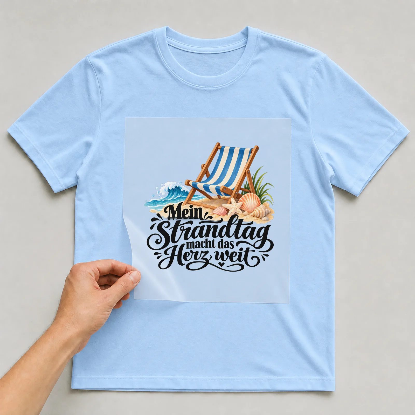 Bügelbild „Mein Strandtag macht das Herz weit“ auf hellblauem T-Shirt, Folie wird aufgelegt