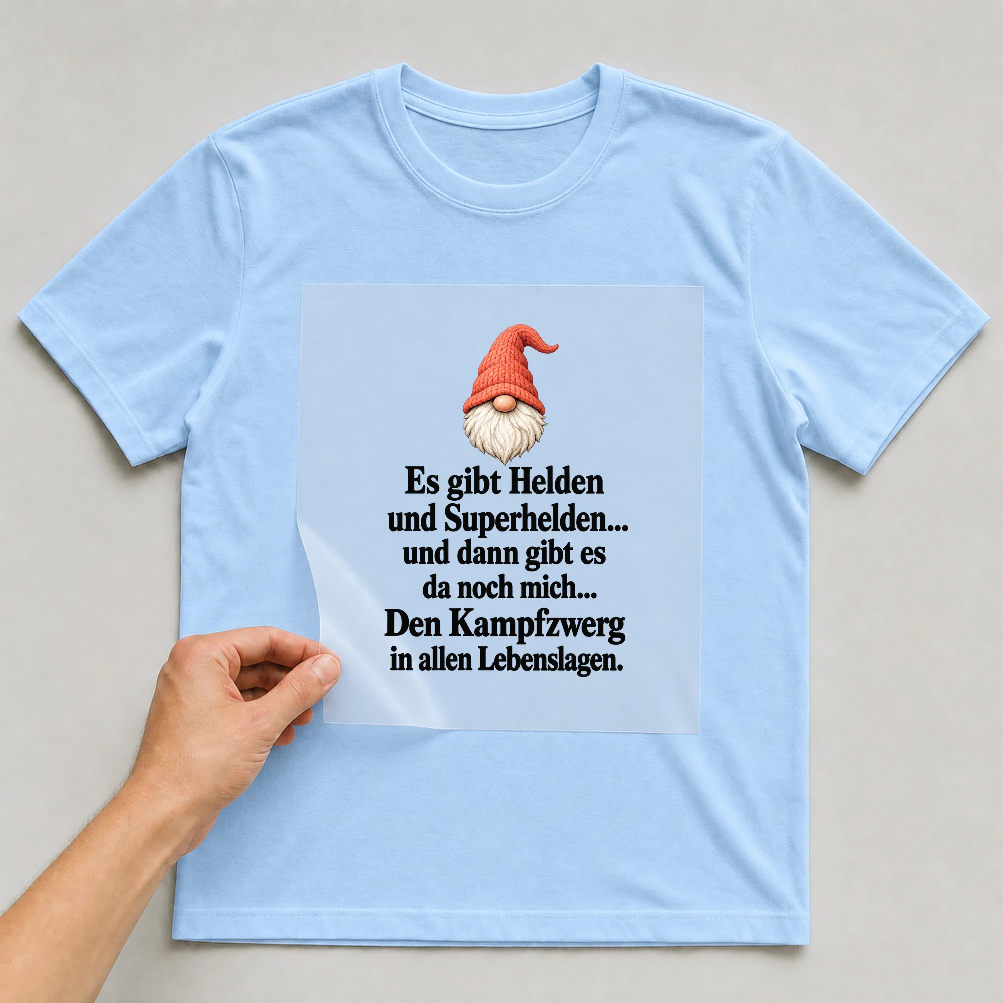 Bügelbild „Es gibt Helden und Superhelden... und dann gibt es da noch mich... Den“ auf babyblauem T-Shirt, Folie wird aufgelegt