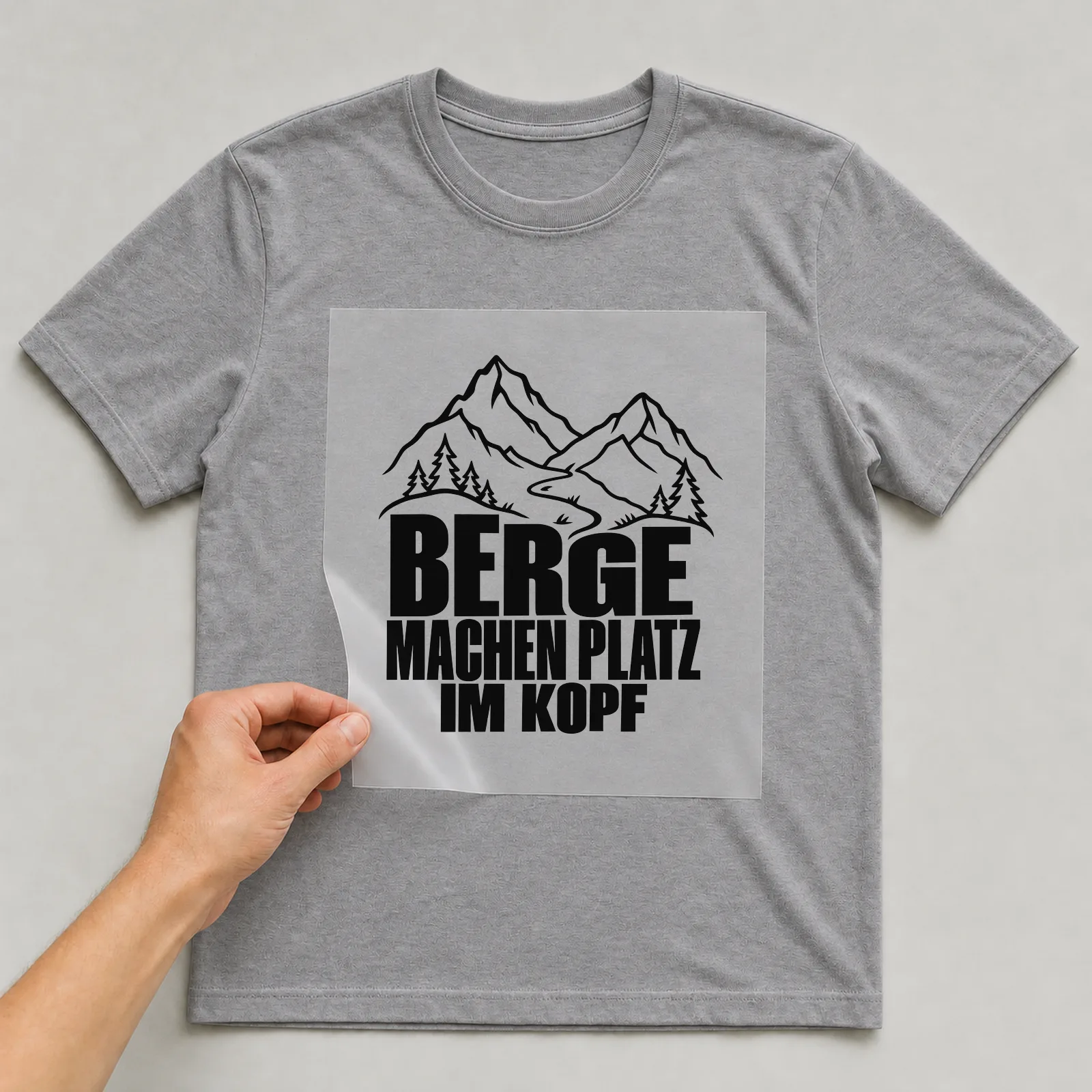 Bügelbild „Berge machen Platz im Kopf“ auf grauem T-Shirt, Folie wird aufgelegt