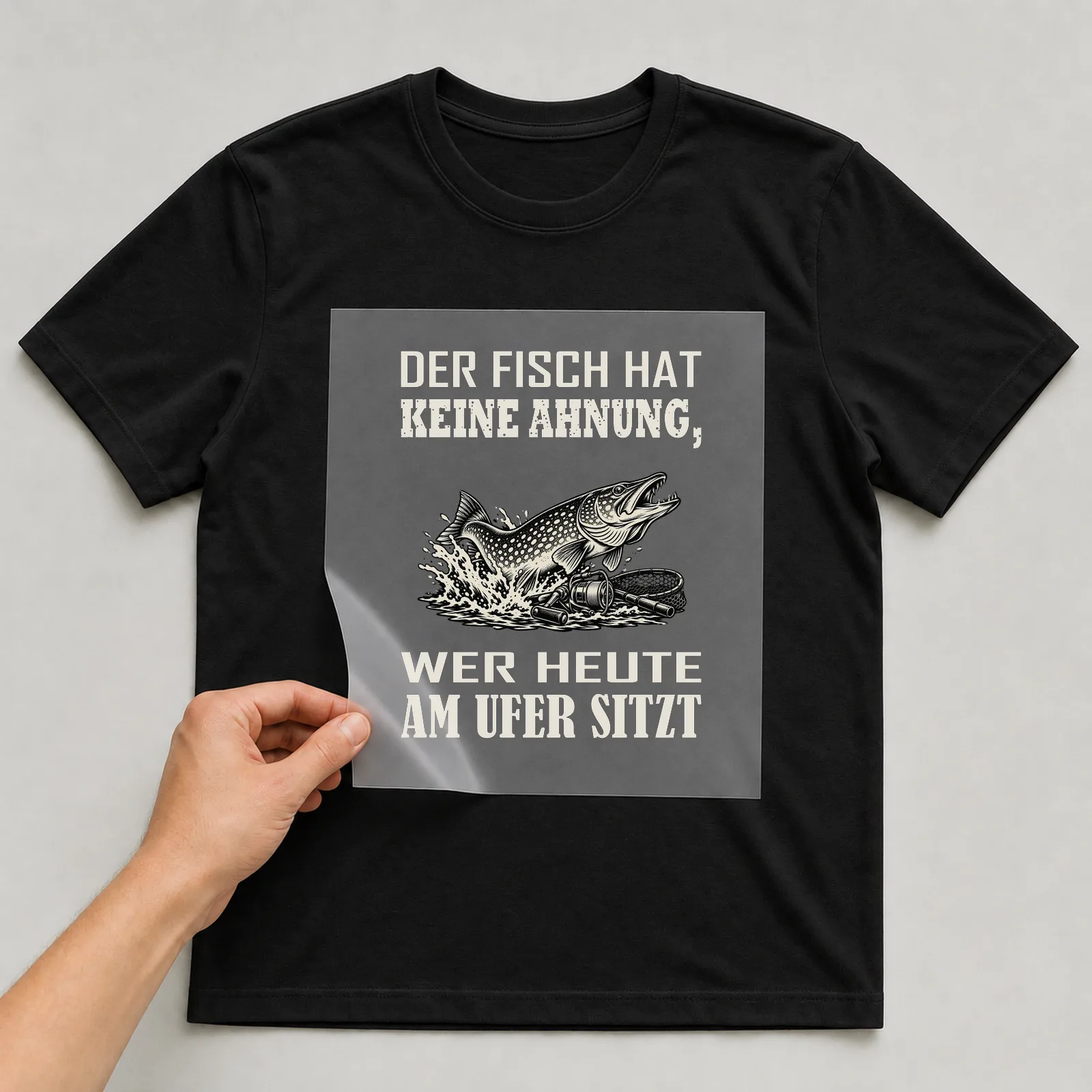 Bügelbild „Der Fisch hat keine Ahnung, wer heute am Ufer sitzt“ auf schwarzem T-Shirt, Folie wird aufgelegt