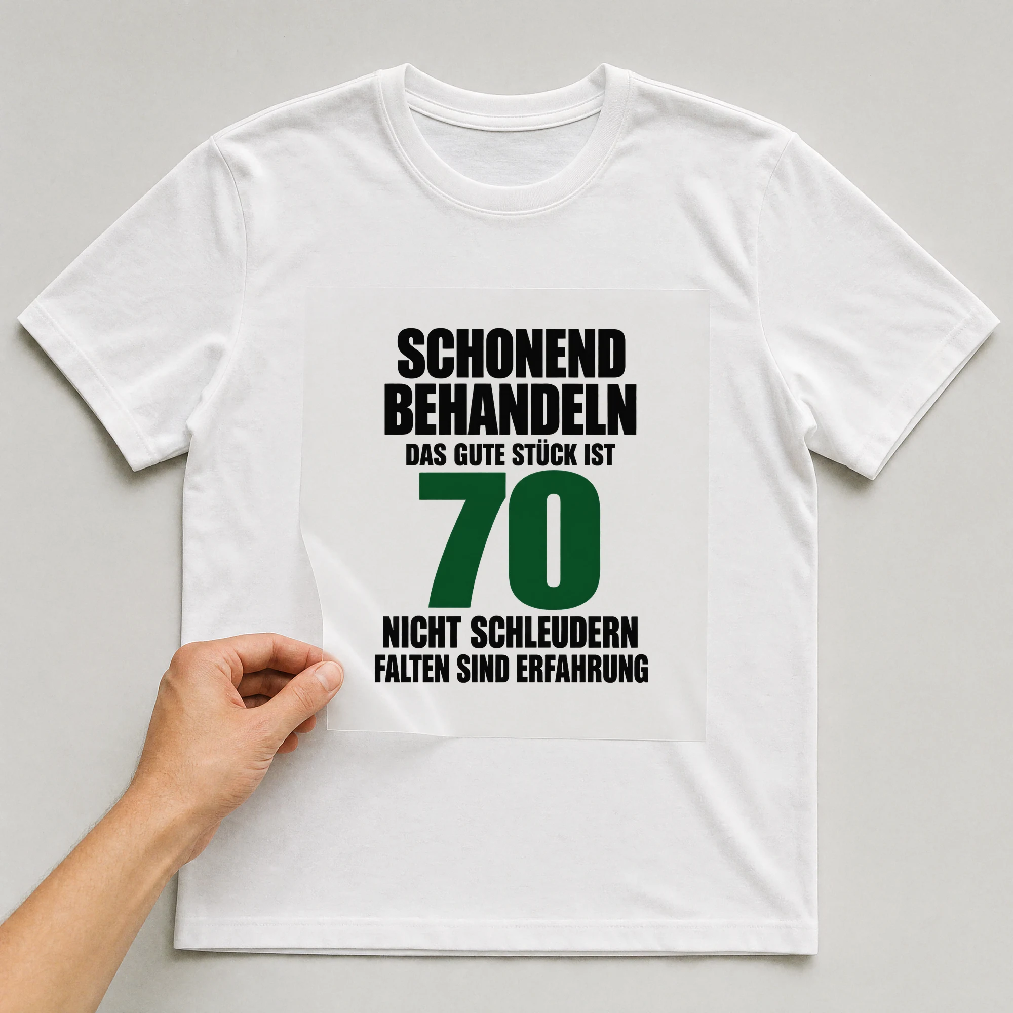 Bügelbild „Schonend behandeln: Das gute Stück ist 70.“ auf weißem T-Shirt, Folie wird aufgelegt