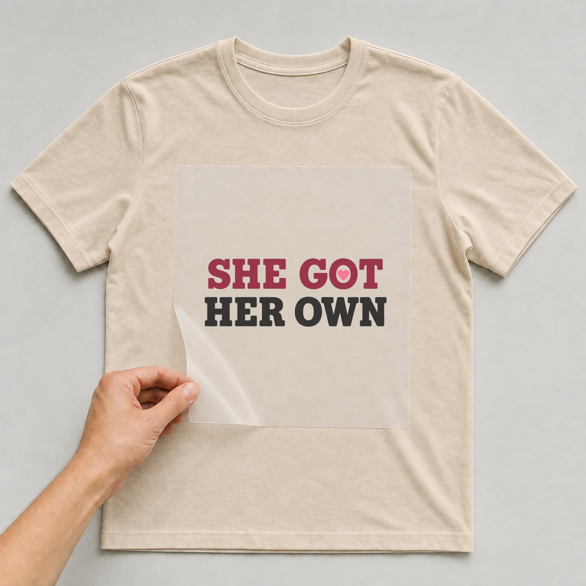 Bügelbild „SHE GOT HER OWN“ auf beigem T-Shirt, Folie wird aufgelegt
