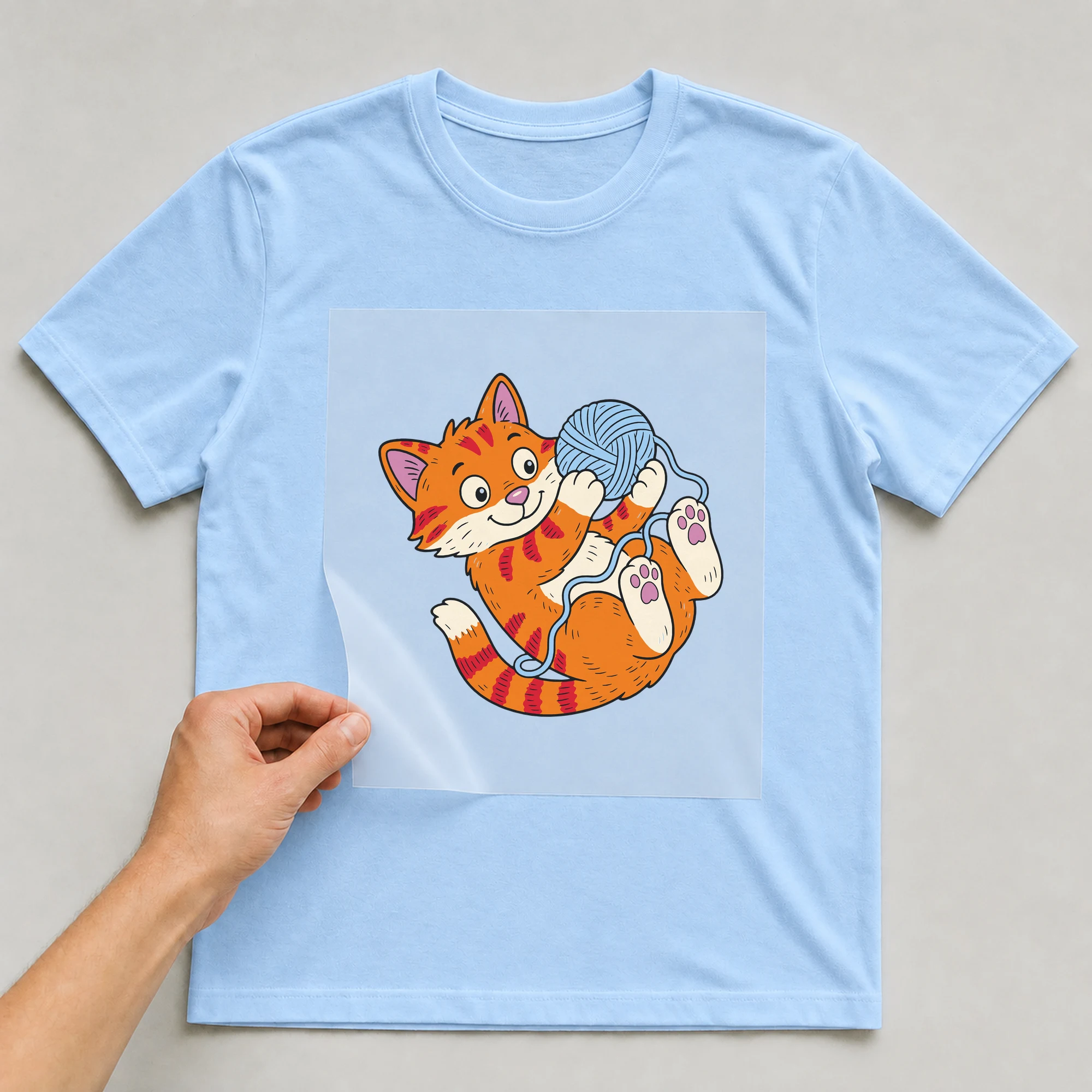 Bügelbild „Katze mit Wollknäuel“ auf babyblauem T-Shirt, Folie wird aufgelegt