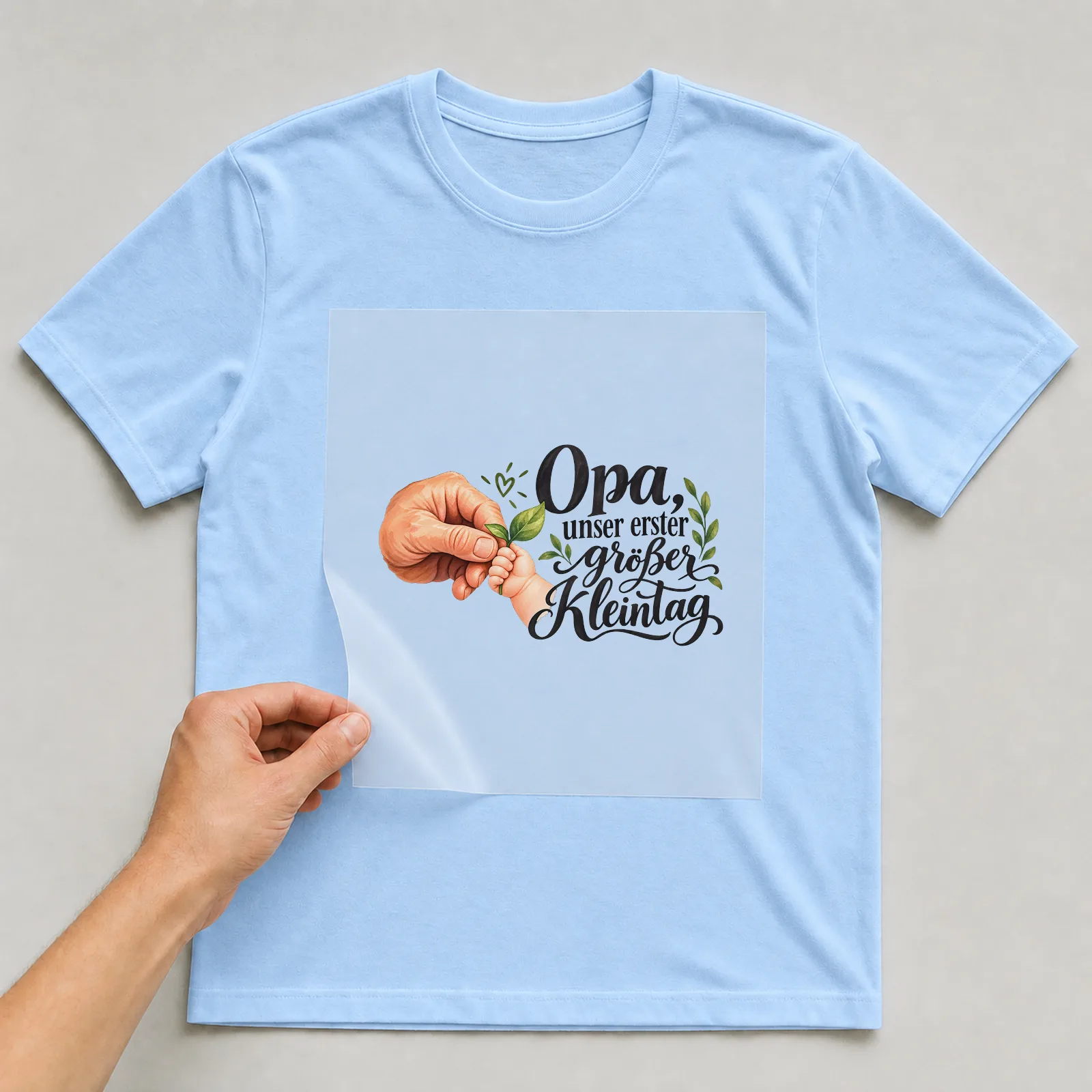 Bügelbild „Opa, unser erster großer Kleintag“ auf hellblauem T-Shirt, Folie wird aufgelegt