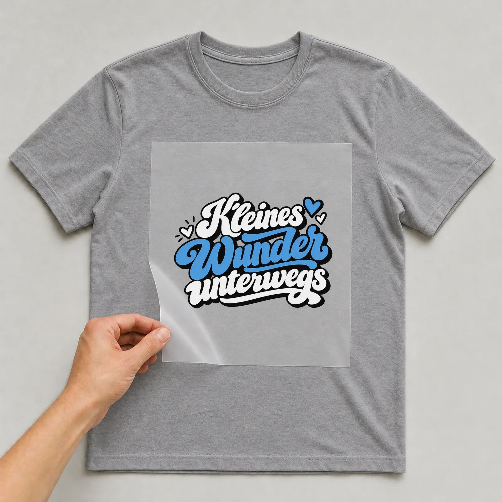 Bügelbild „Kleines Wunder unterwegs“ auf grauem T-Shirt, Folie wird aufgelegt
