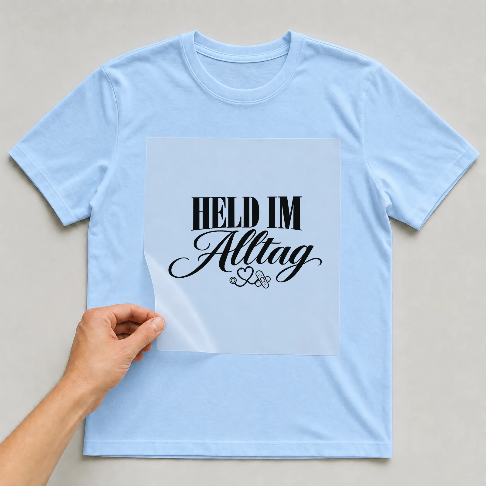 Bügelbild „Held im Alltag“ auf hellblauem T-Shirt, Folie wird aufgelegt