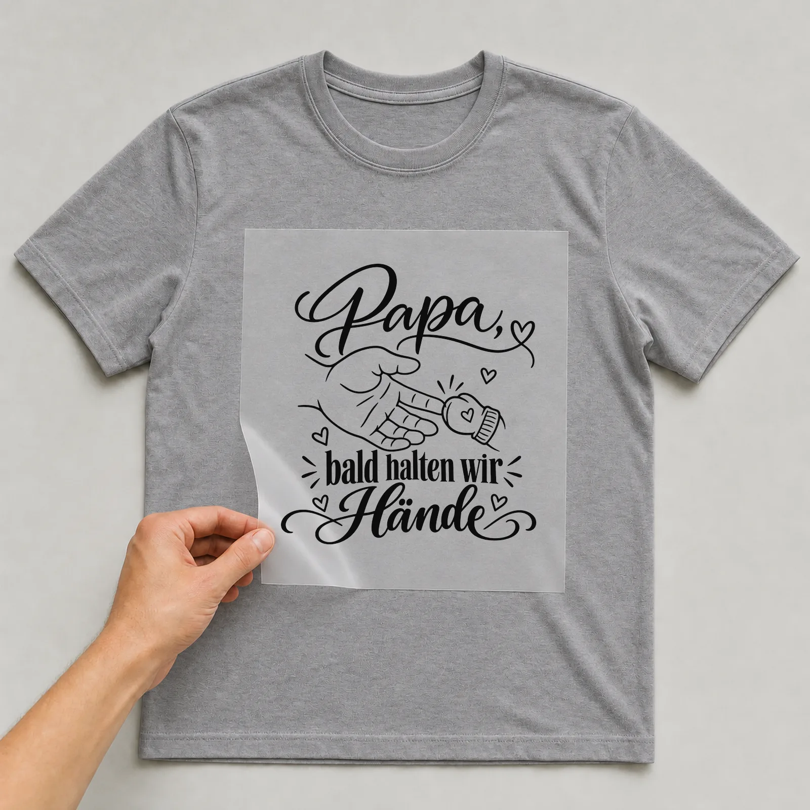 Bügelbild „Papa, bald halten wir Hände“ auf grauem T-Shirt, Folie wird aufgelegt