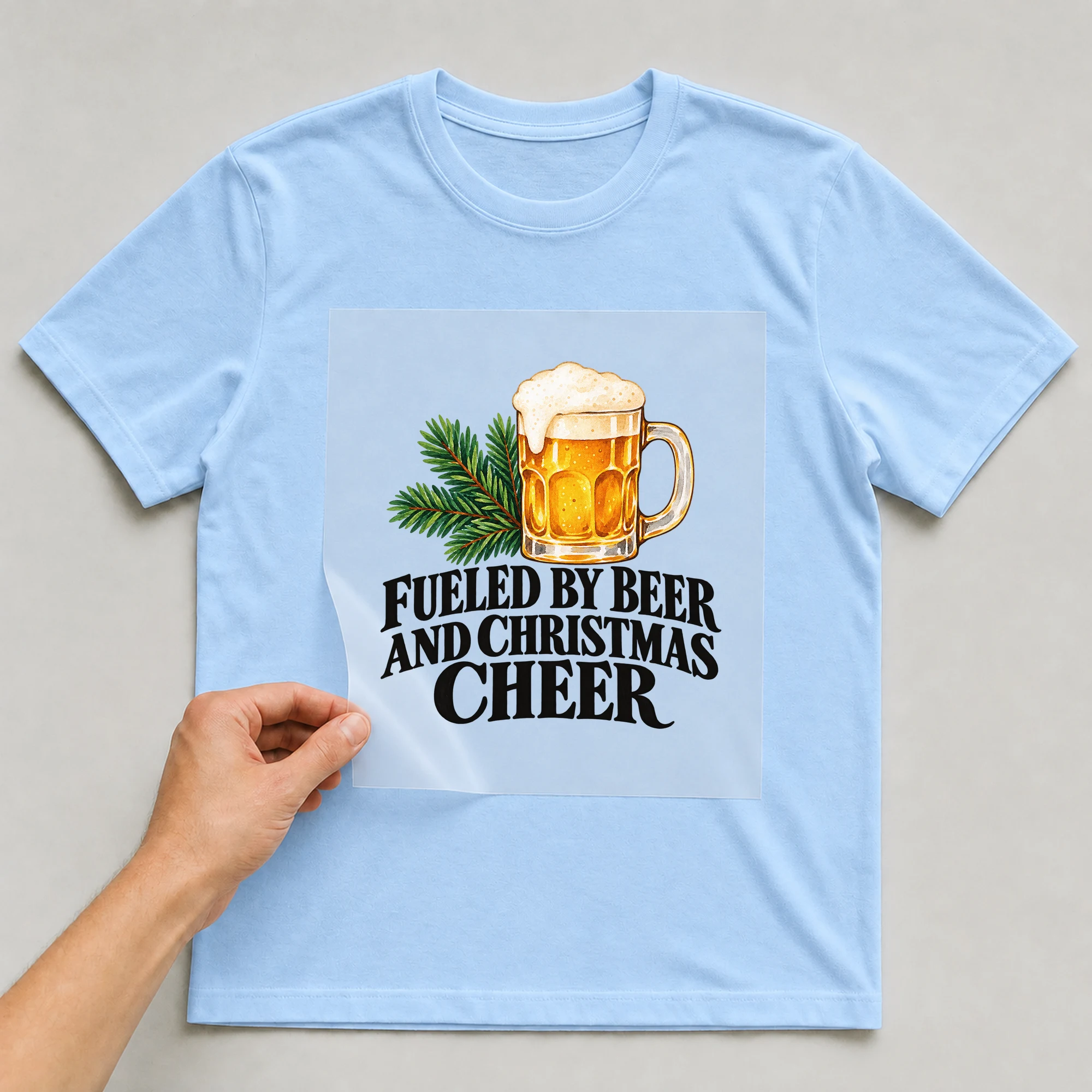 Bügelbild „Fueled By Beer And Christmas Cheer“ auf babyblauem T-Shirt, Folie wird aufgelegt