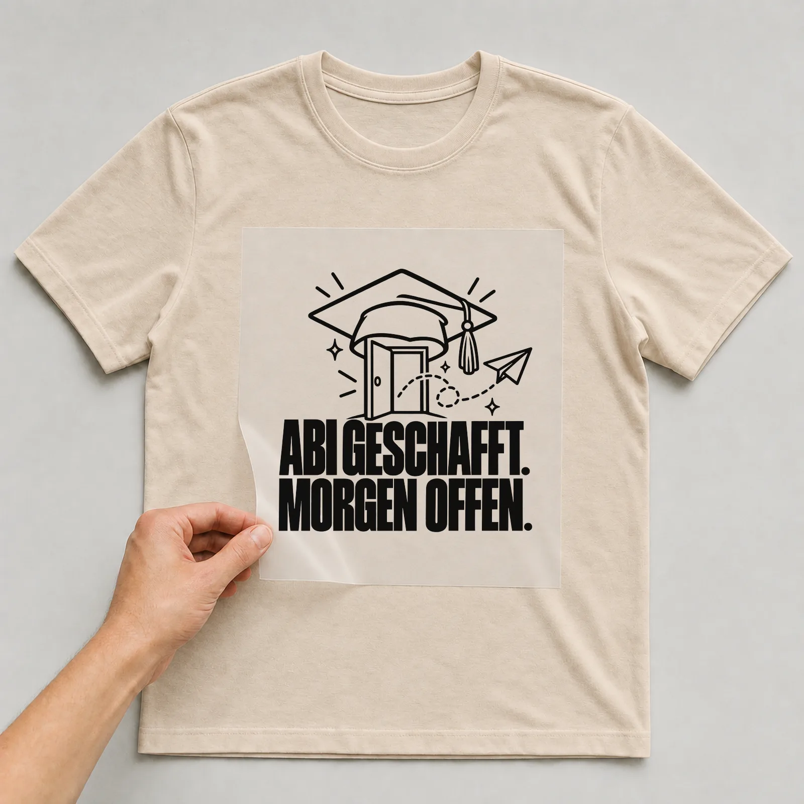 Bügelbild „Abi geschafft. Morgen offen.“ auf beigem T-Shirt, Folie wird aufgelegt