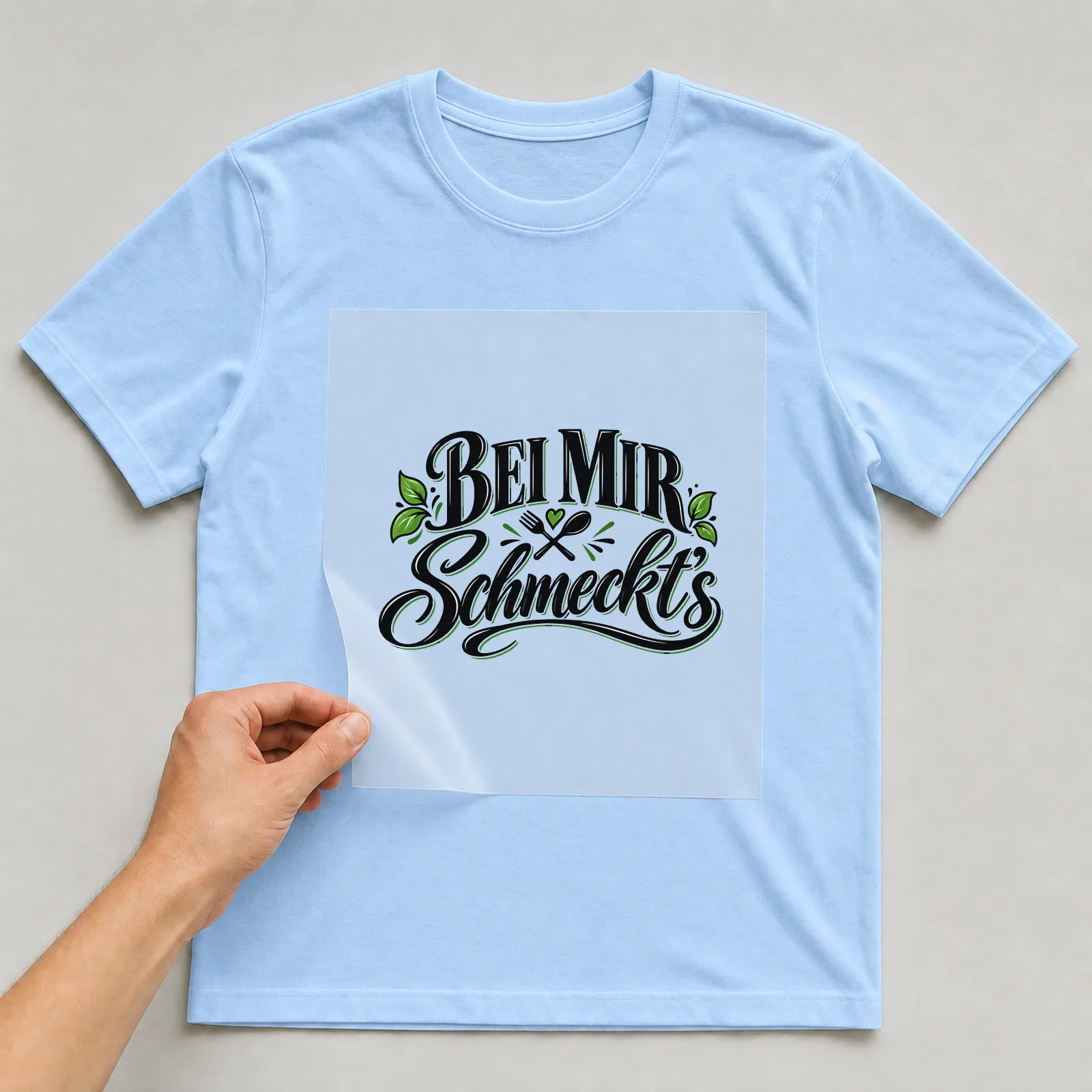 Bügelbild „Bei mir schmeckt’s“ auf hellblauem T-Shirt, Folie wird aufgelegt