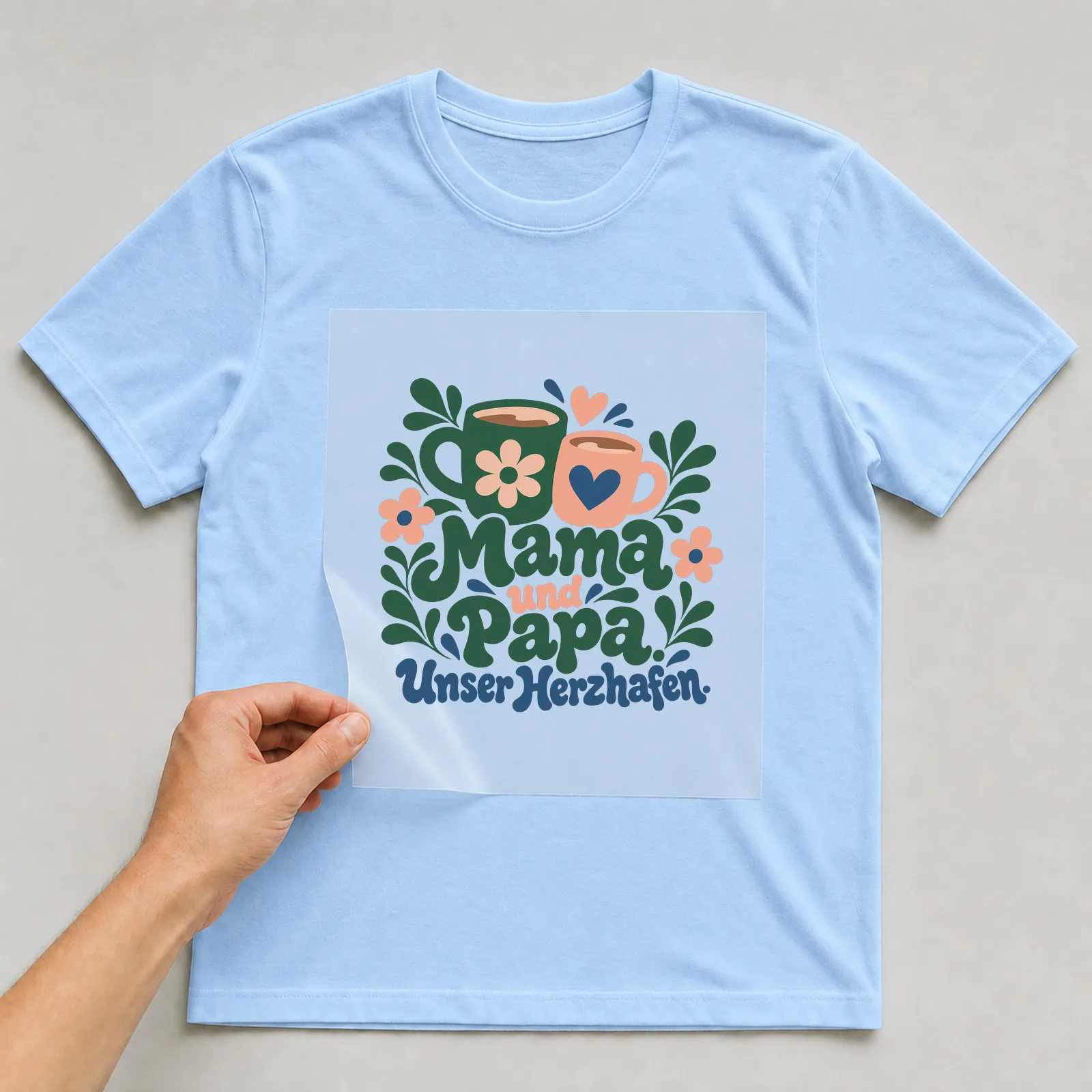 Bügelbild „Mama und Papa. Unser Herzhafen.“ auf hellblauem T-Shirt, Folie wird aufgelegt