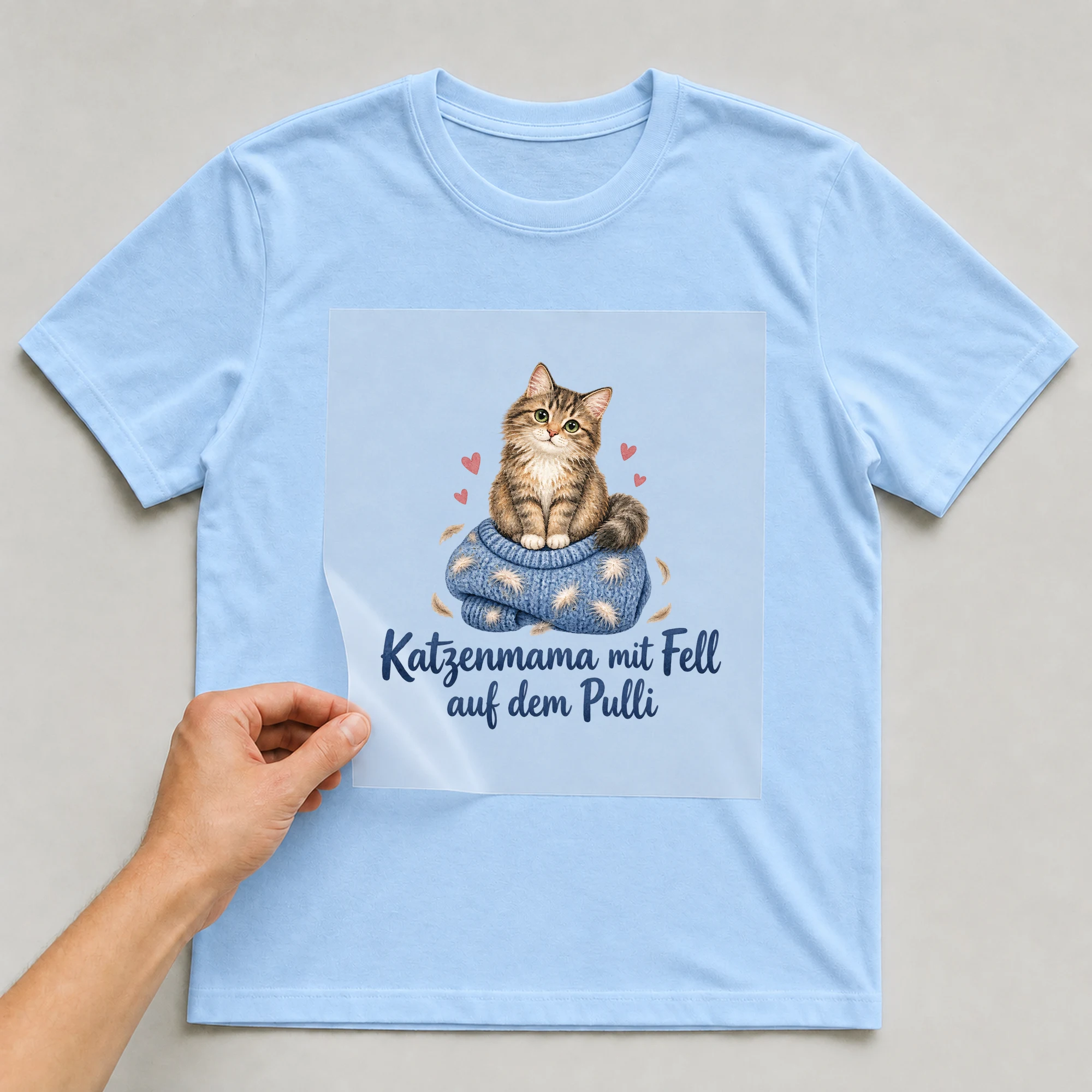 Bügelbild „Katzenmama mit Fell auf dem Pulli“ auf babyblauem T-Shirt, Folie wird aufgelegt