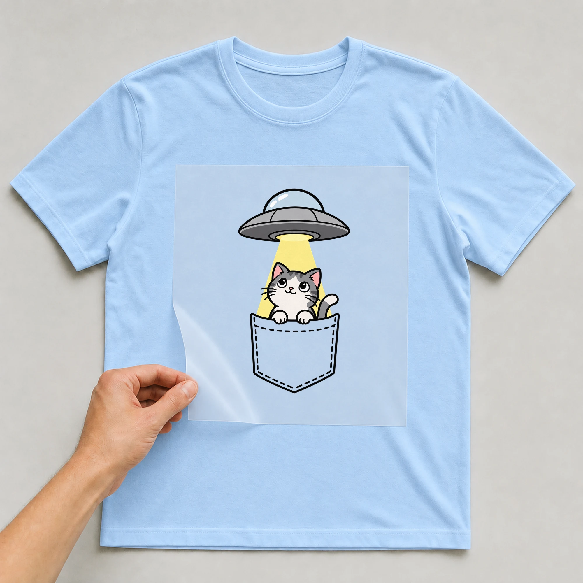 Bügelbild „Brusttasche Katze Aliens“ auf babyblauem T-Shirt, Folie wird aufgelegt