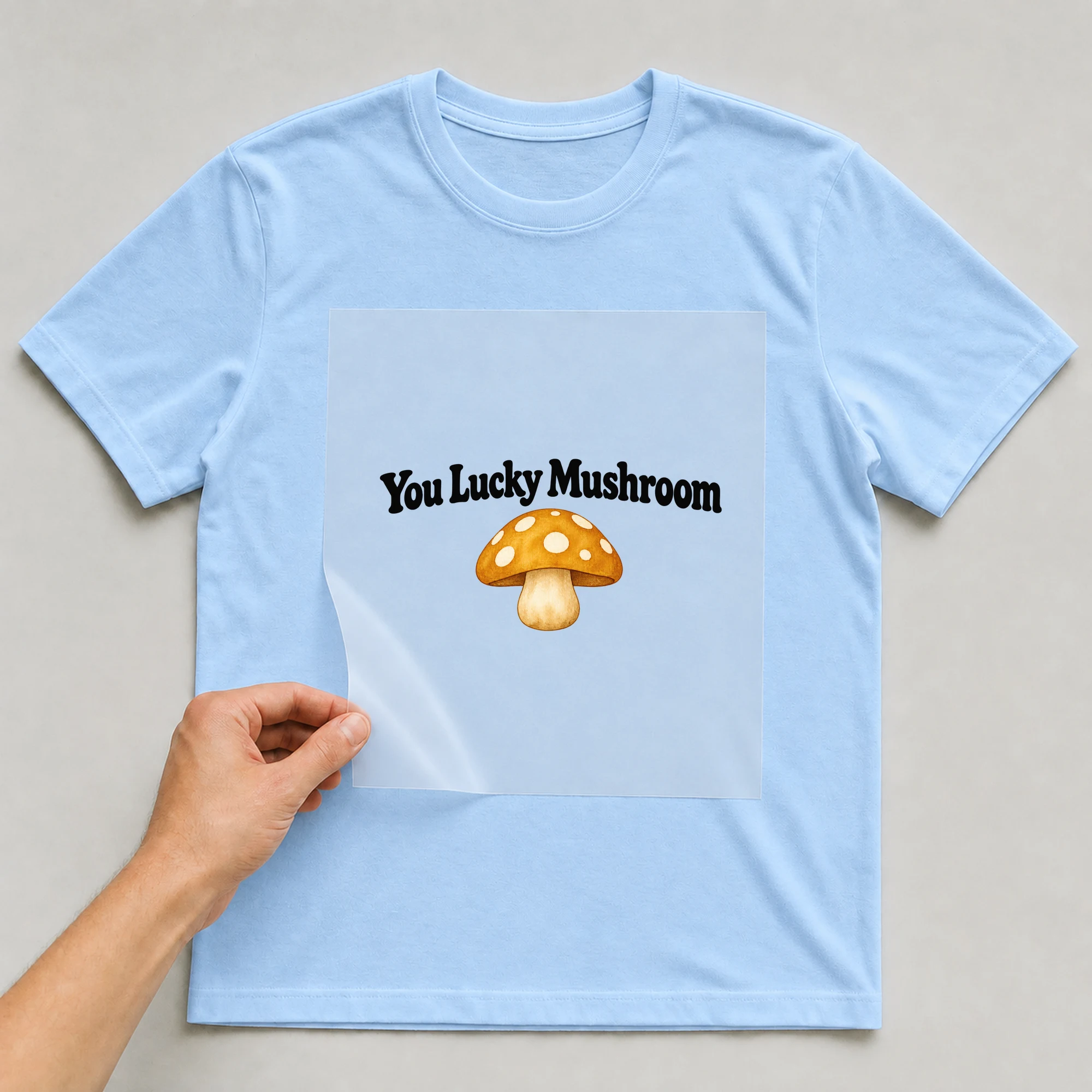 Bügelbild „You Lucky Mushroom“ auf babyblauem T-Shirt, Folie wird aufgelegt