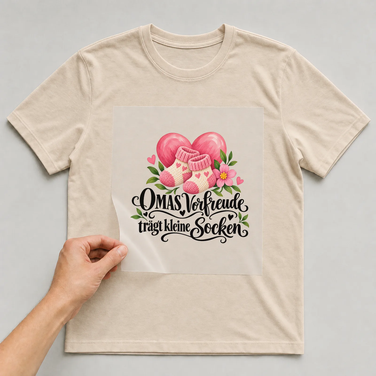 Bügelbild „OMAS Vorfreude trägt kleine Socken“ auf beigem T-Shirt, Folie wird aufgelegt