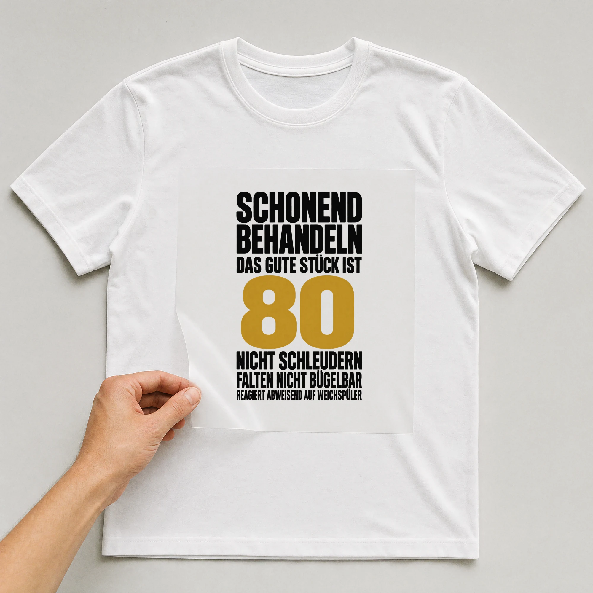 Bügelbild „Schonend behandeln das gute Stück ist 80 nicht schleudern falten nicht“ auf weißem T-Shirt, Folie wird aufgelegt