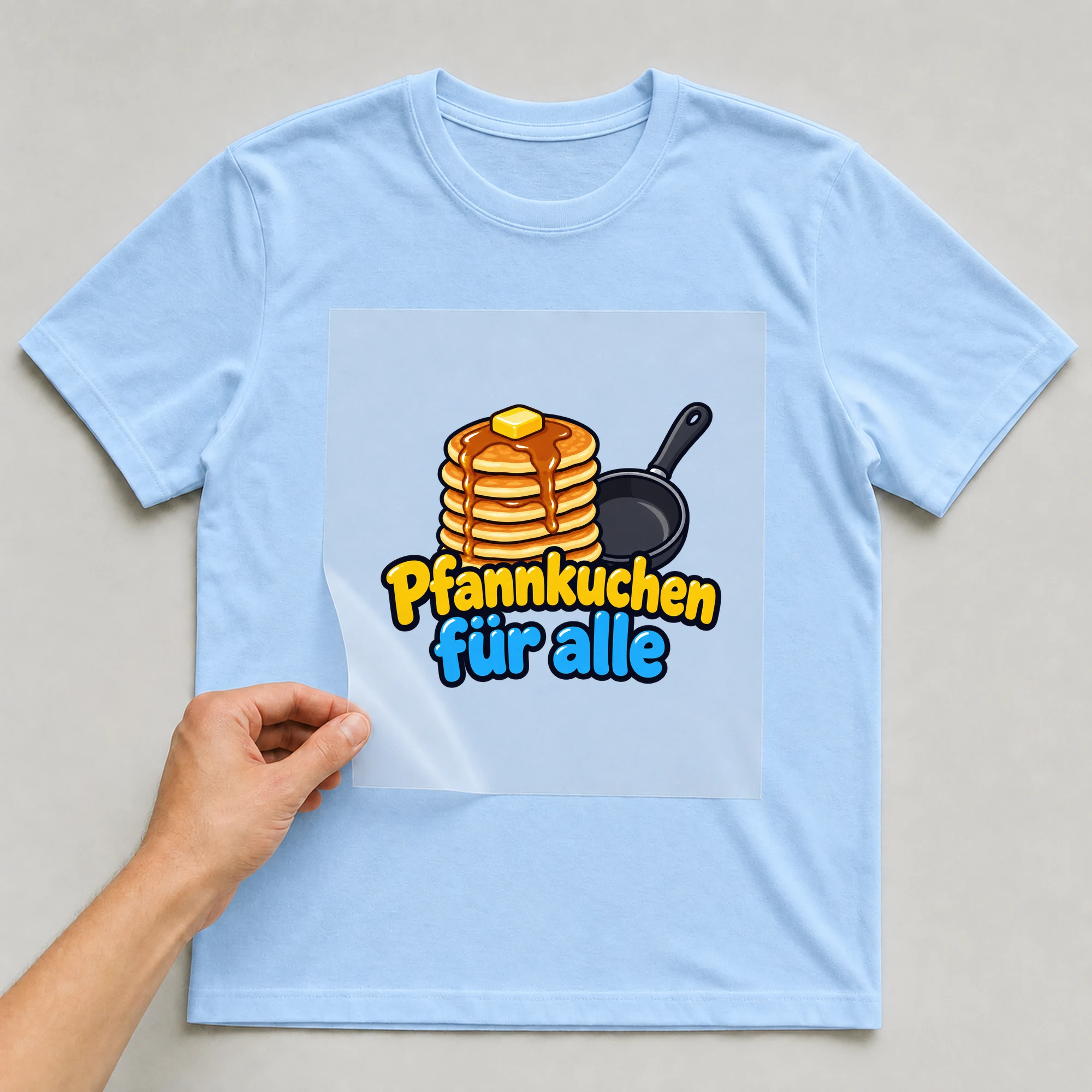Bügelbild „Pfannkuchen für alle“ auf babyblauem T-Shirt, Folie wird aufgelegt