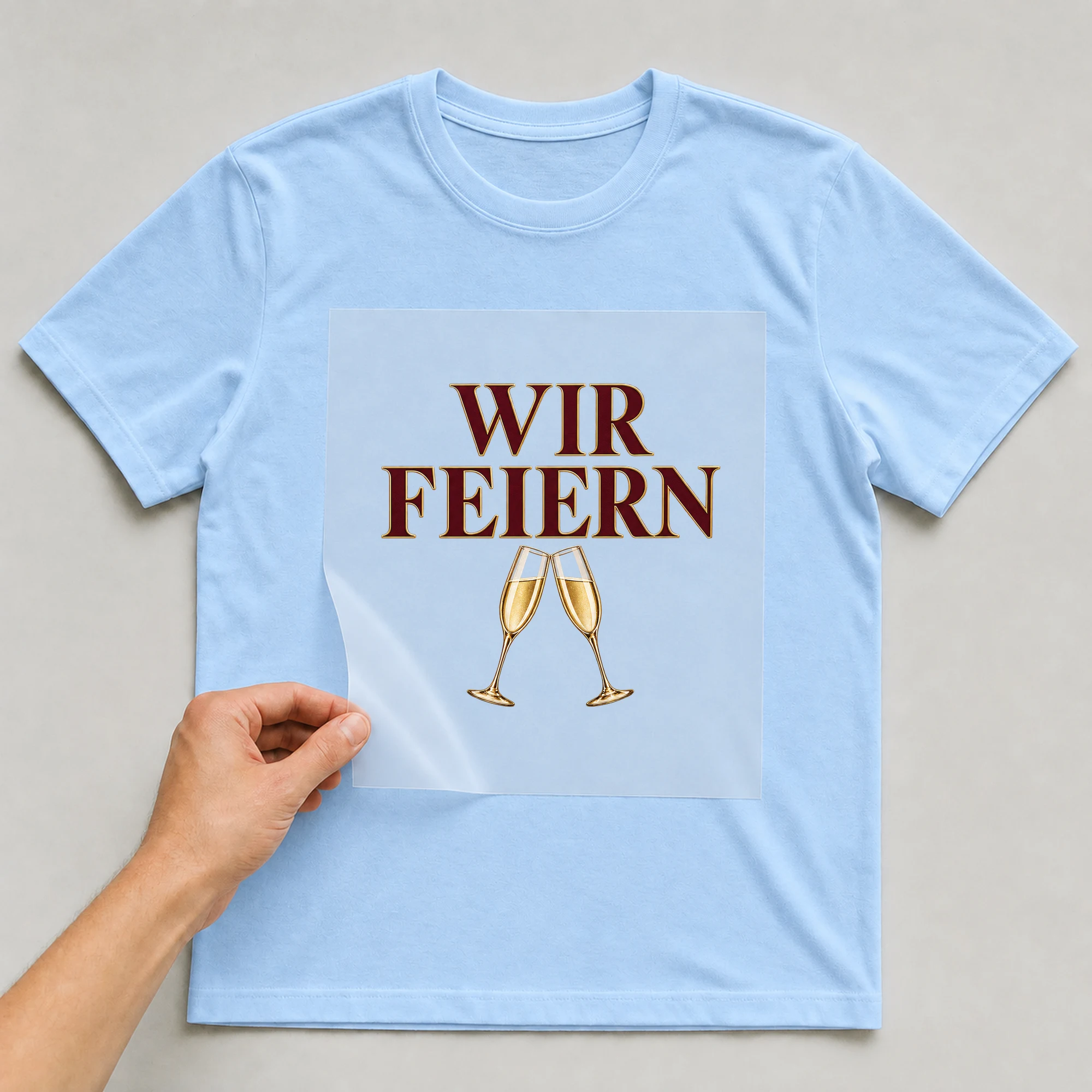 Bügelbild „Wir Feiern“ auf babyblauem T-Shirt, Folie wird aufgelegt