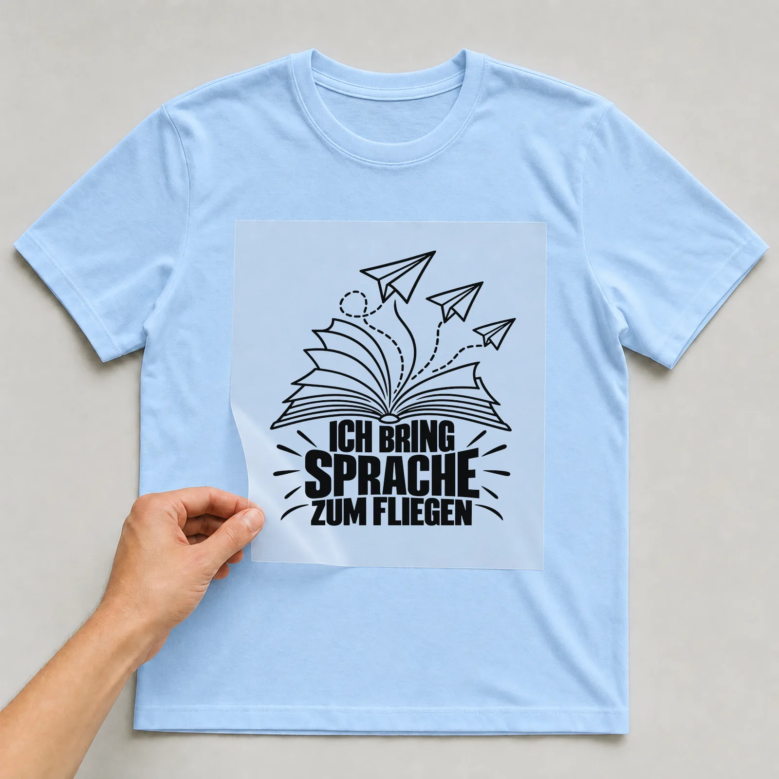 Bügelbild „Ich bring Sprache zum Fliegen“ auf hellblauem T-Shirt, Folie wird aufgelegt