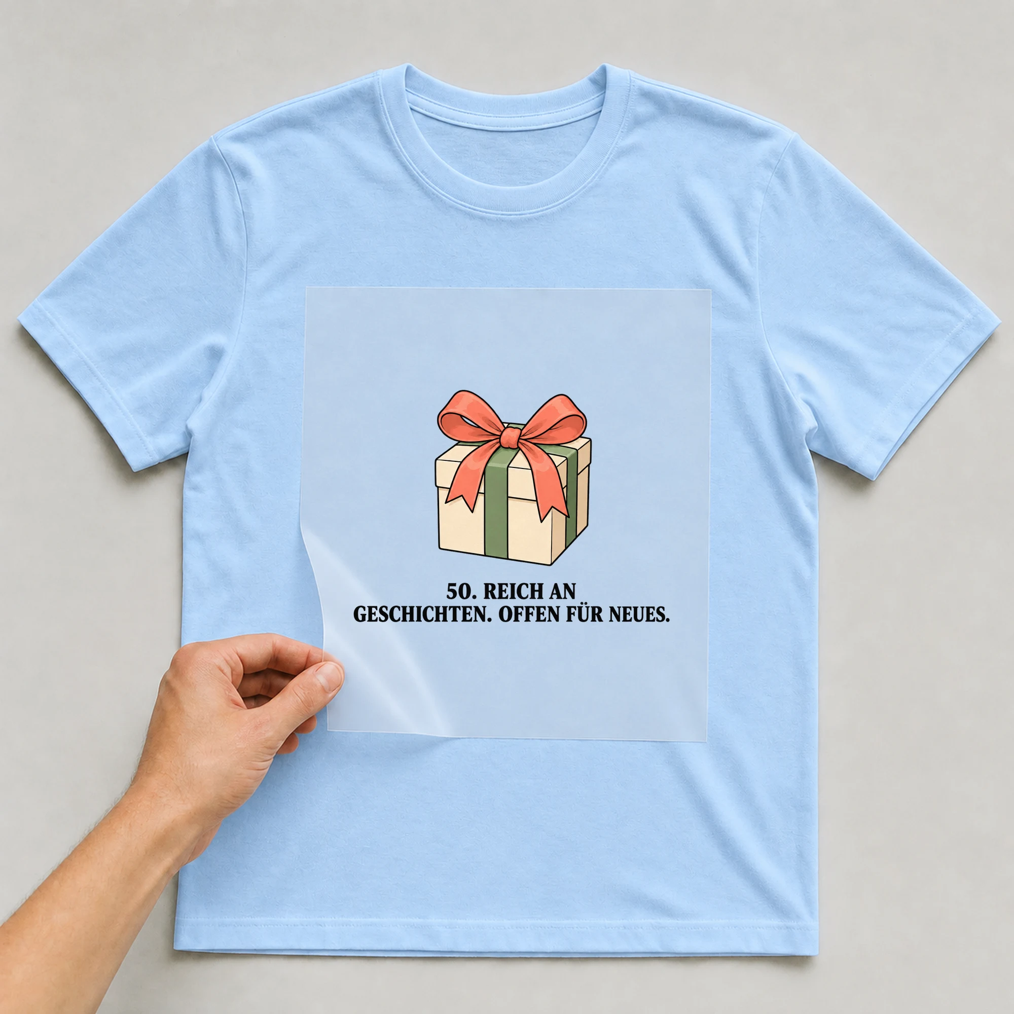 Bügelbild „50. REICH AN GESCHICHTEN. OFFEN FÜR NEUES.“ auf babyblauem T-Shirt, Folie wird aufgelegt