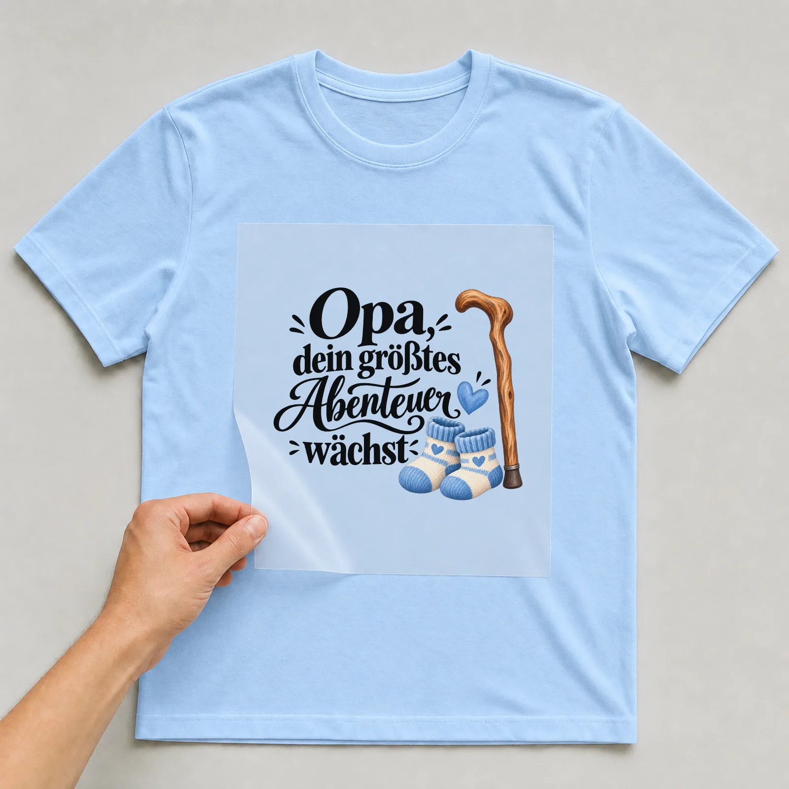 Bügelbild „Opa, dein größtes Abenteuer wächst“ auf hellblauem T-Shirt, Folie wird aufgelegt