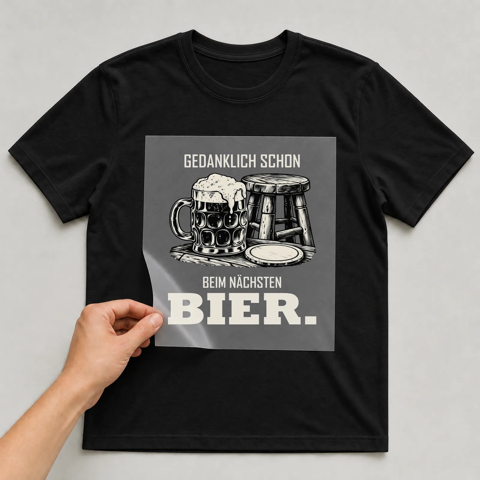 Bügelbild „Gedanklich schon beim nächsten Bier“ auf schwarzem T-Shirt, Folie wird aufgelegt