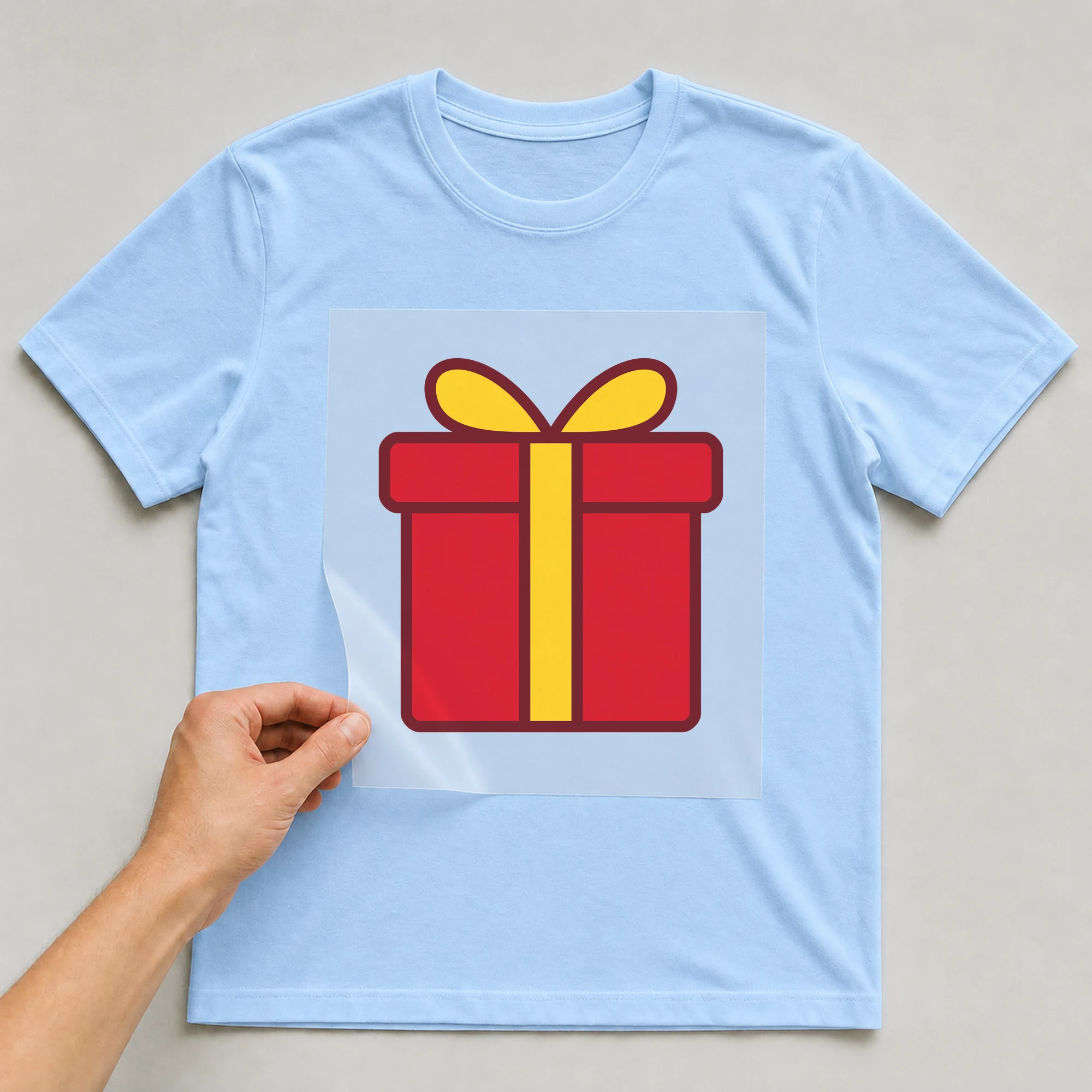 Bügelbild „Geschenk mit Schleife“ auf babyblauem T-Shirt, Folie wird aufgelegt