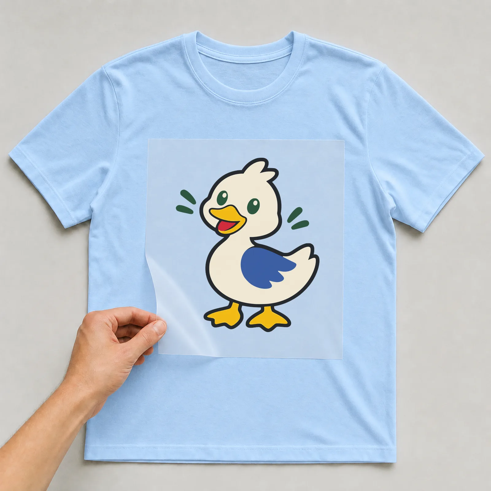 Bügelbild Ente im Comic-Stil auf hellblauem T-Shirt, Folie wird aufgelegt