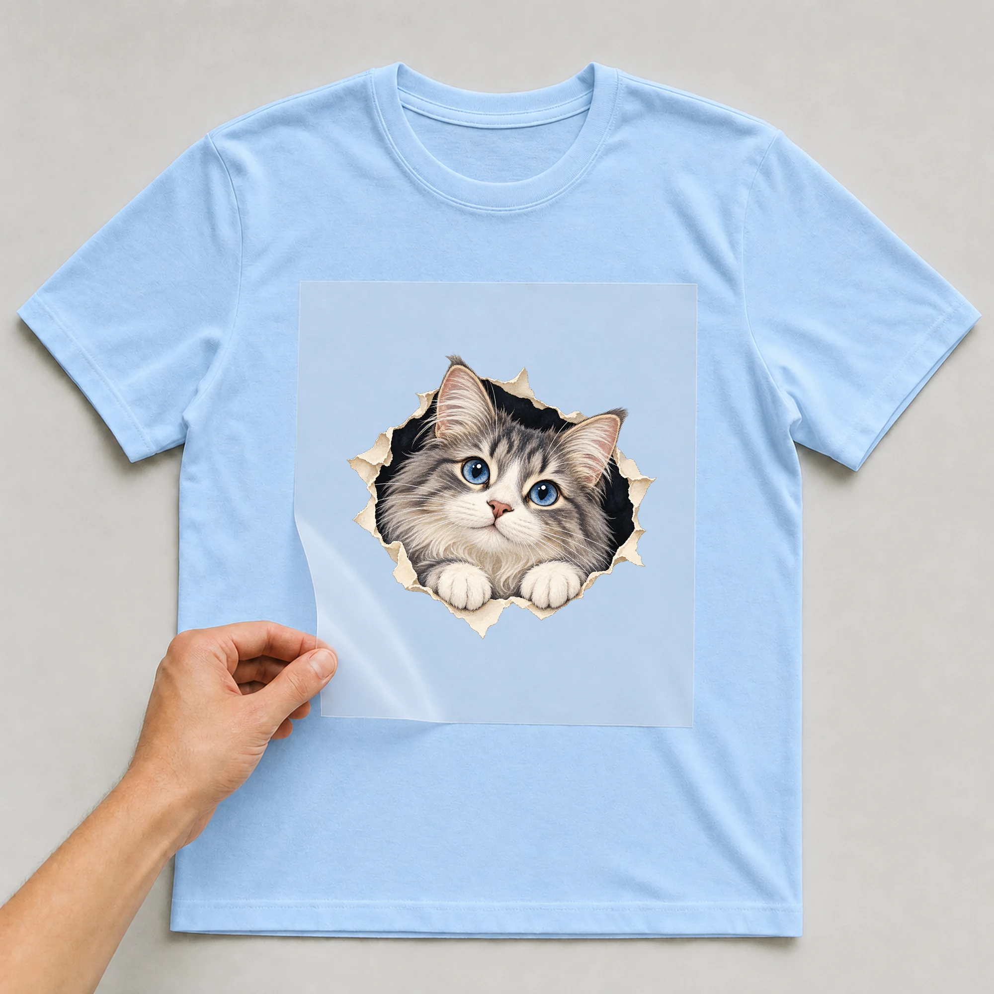 Bügelbild „Katze Schaut Durch Ein Guckloch“ auf babyblauem T-Shirt, Folie wird aufgelegt