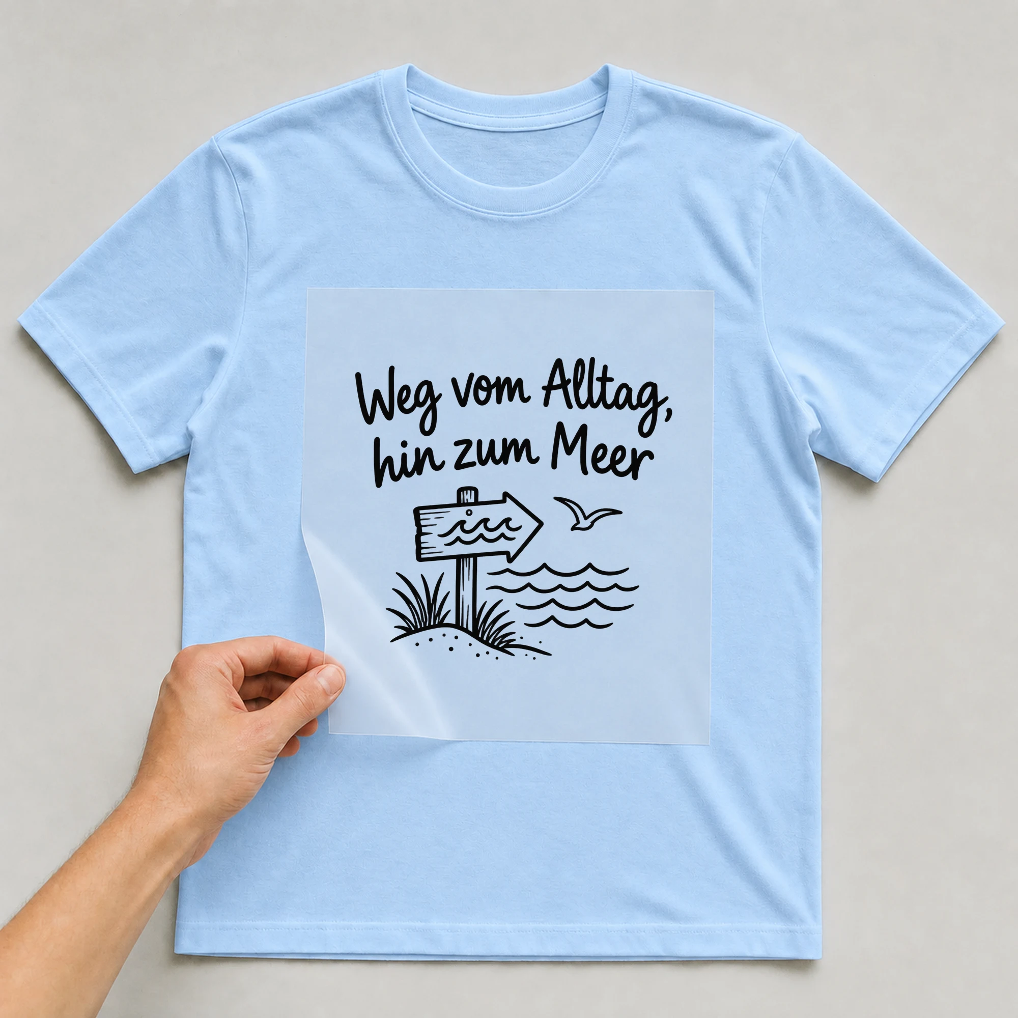 Bügelbild „Weg vom Alltag, hin zum Meer“ auf babyblauem T-Shirt, Folie wird aufgelegt