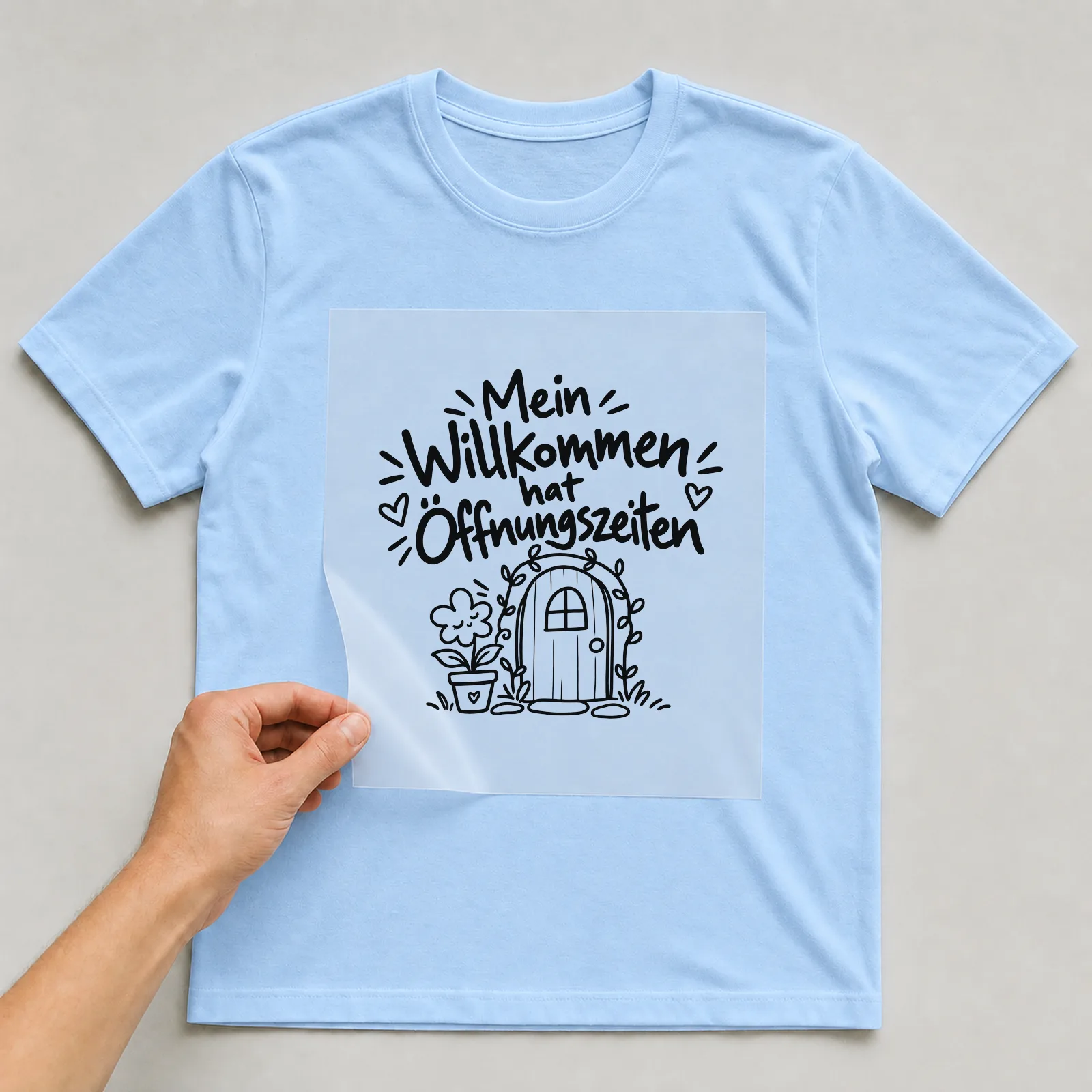 Bügelbild „Mein Willkommen hat Öffnungszeiten“ auf hellblauem T-Shirt, Folie wird aufgelegt
