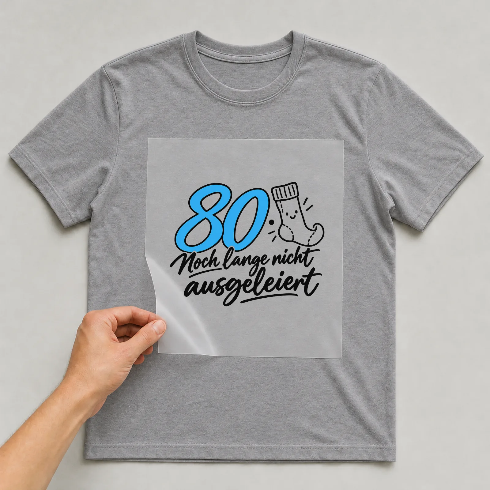 Bügelbild „80 · Noch lange nicht ausgeleiert“ auf grauem T-Shirt, Folie wird aufgelegt
