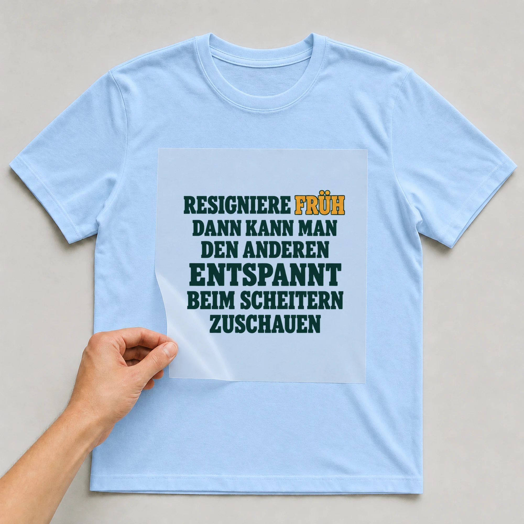 Bügelbild „RESIGNIERE FRÜH DANN KANN MAN DEN ANDEREN ENTSPANNT BEIM SCHEITERN ZUSCHAUEN“ auf babyblauem T-Shirt, Folie wird aufgelegt