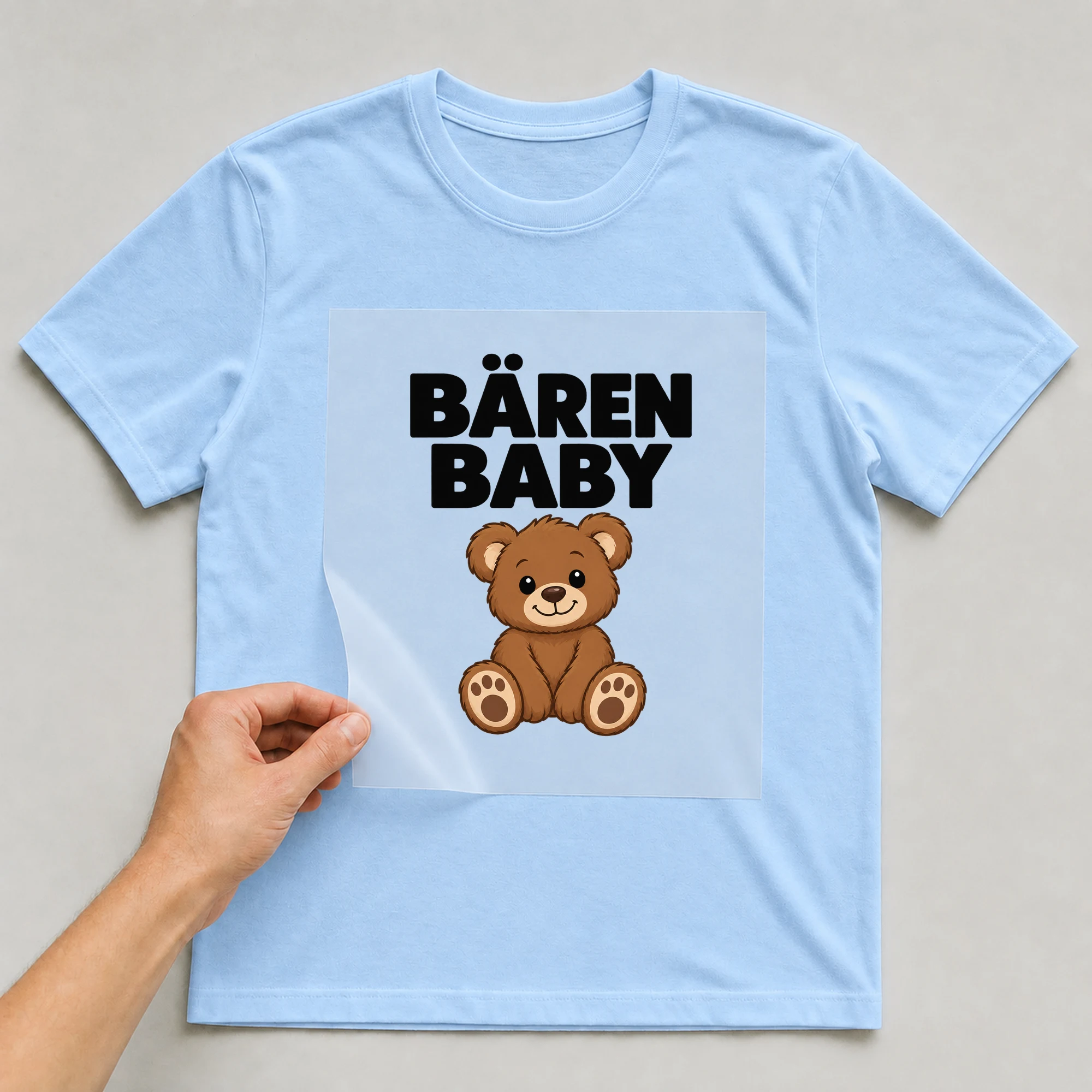 Bügelbild „Bärenbaby“ auf babyblauem T-Shirt, Folie wird aufgelegt