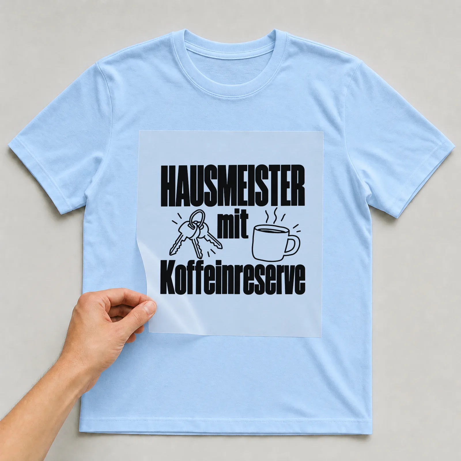 Bügelbild „Hausmeister mit Koffeinreserve“ auf hellblauem T-Shirt, Folie wird aufgelegt