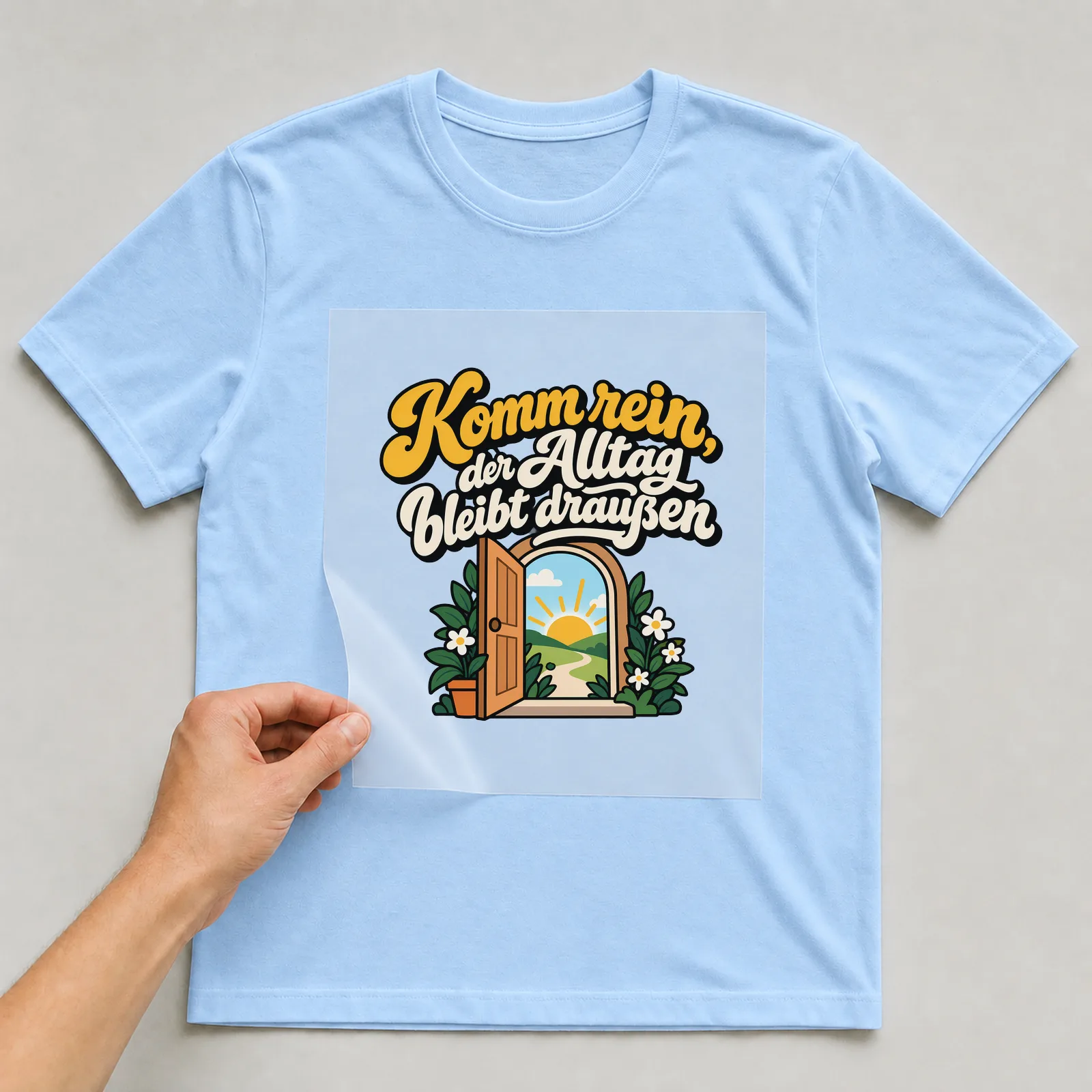 Bügelbild „Komm rein, der Alltag bleibt draußen“ auf hellblauem T-Shirt, Folie wird aufgelegt