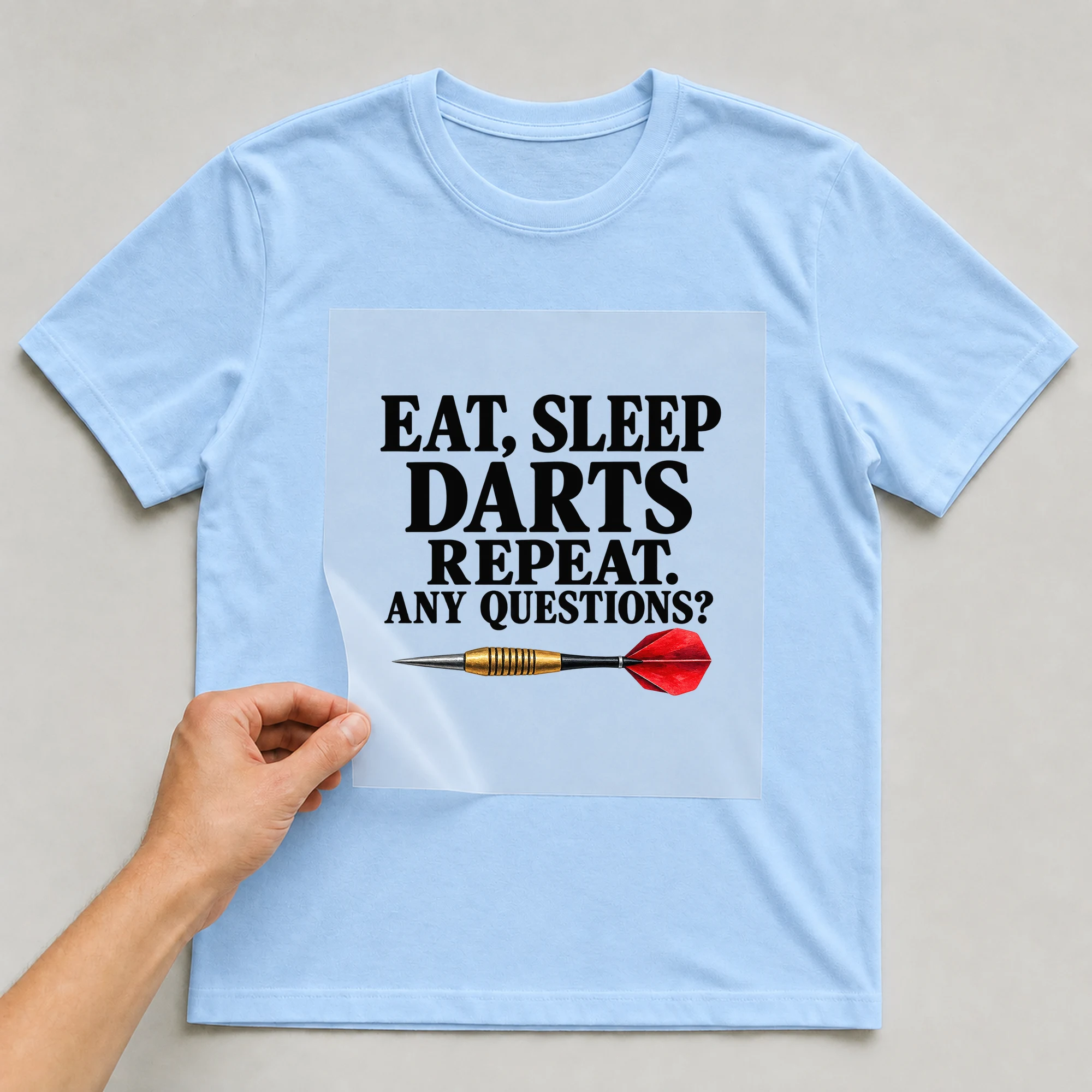 Bügelbild „Eat, Sleep Darts Repeat. Any Questions?“ auf babyblauem T-Shirt, Folie wird aufgelegt
