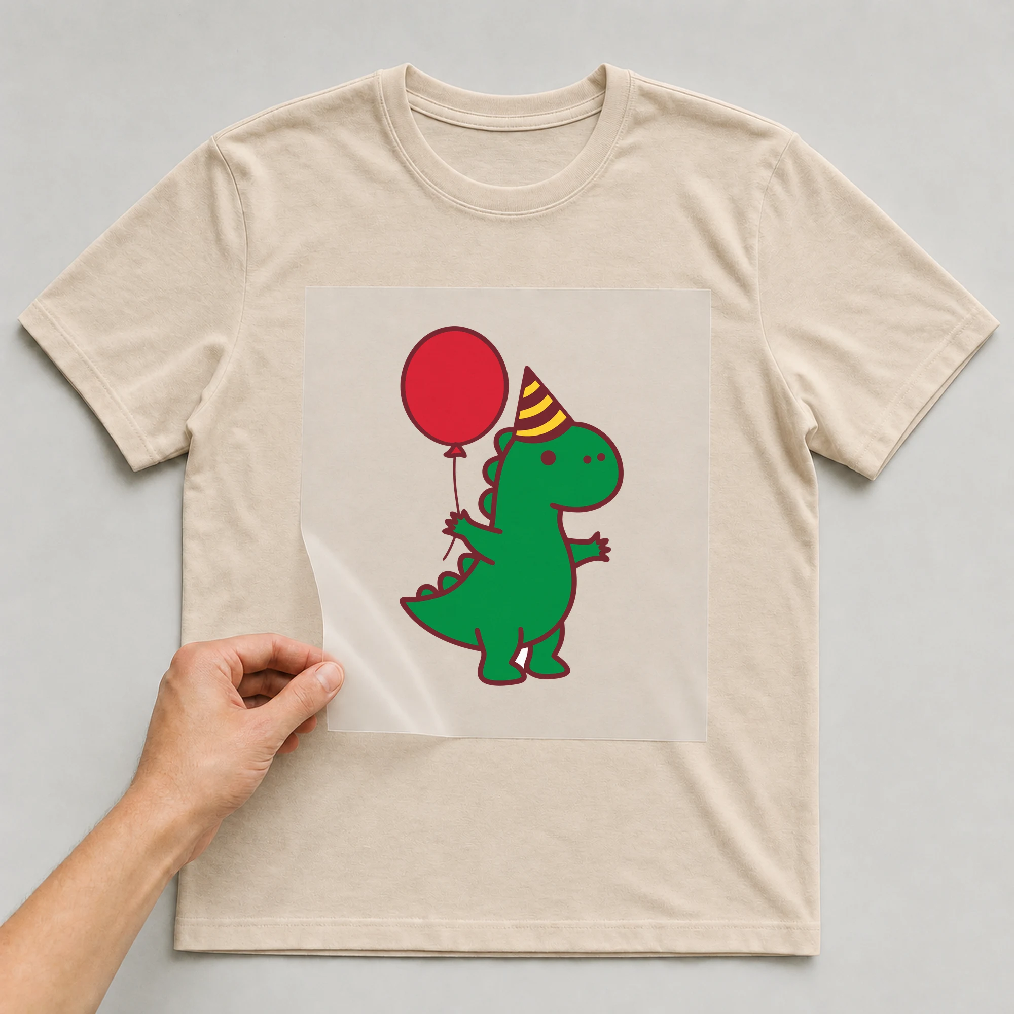 Bügelbild „Dino mit Partyhut“ auf beigem T-Shirt, Folie wird aufgelegt