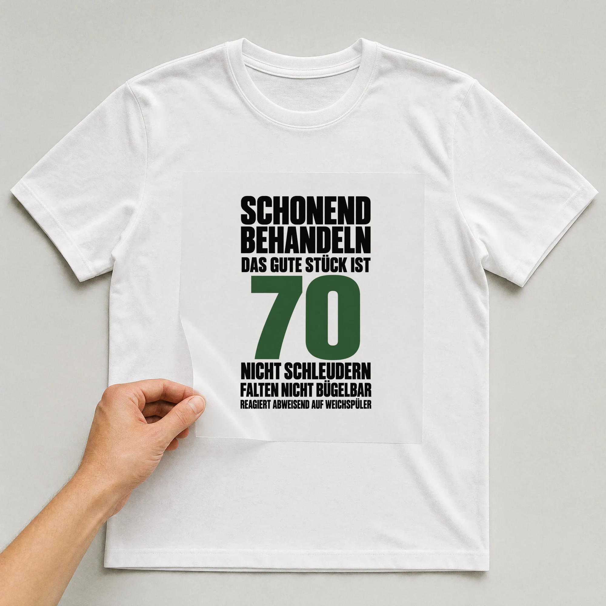 Bügelbild „Schonend behandeln das gute Stück ist 70 nicht schleudern falten nicht“ auf weißem T-Shirt, Folie wird aufgelegt