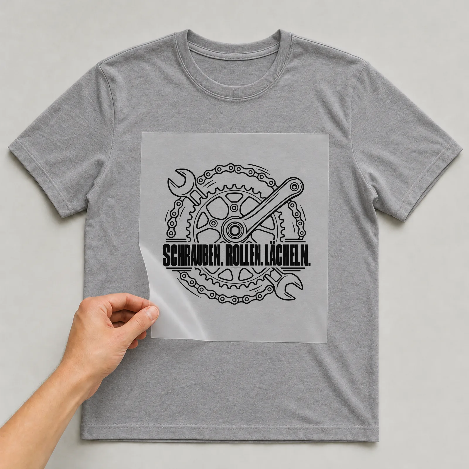 Bügelbild „Schrauben. Rollen. Lächeln.“ auf grauem T-Shirt, Folie wird aufgelegt