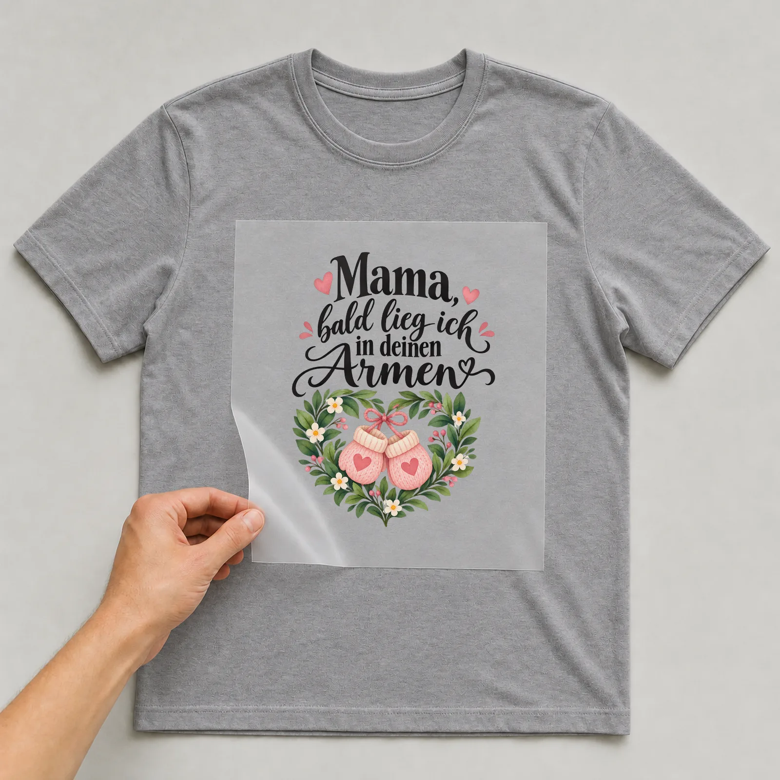 Bügelbild „Mama, bald lieg ich in deinen Armen“ auf grauem T-Shirt, Folie wird aufgelegt
