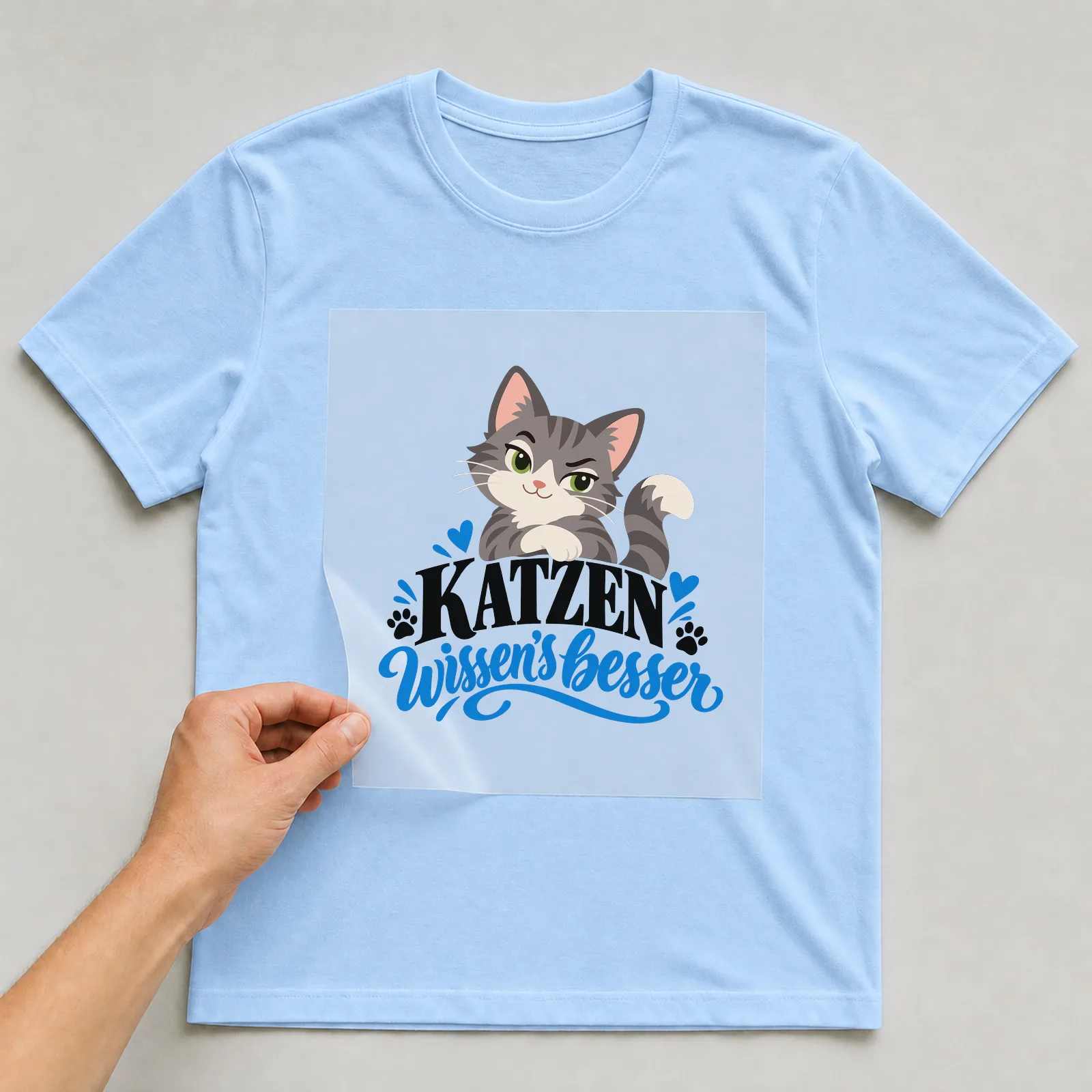 Bügelbild „Katzen wissen’s besser“ auf hellblauem T-Shirt, Folie wird aufgelegt