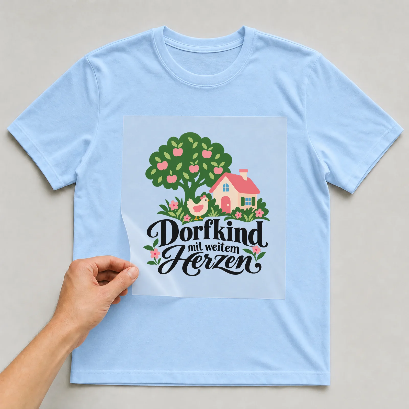 Bügelbild „Dorfkind mit weitem Herzen“ auf hellblauem T-Shirt, Folie wird aufgelegt